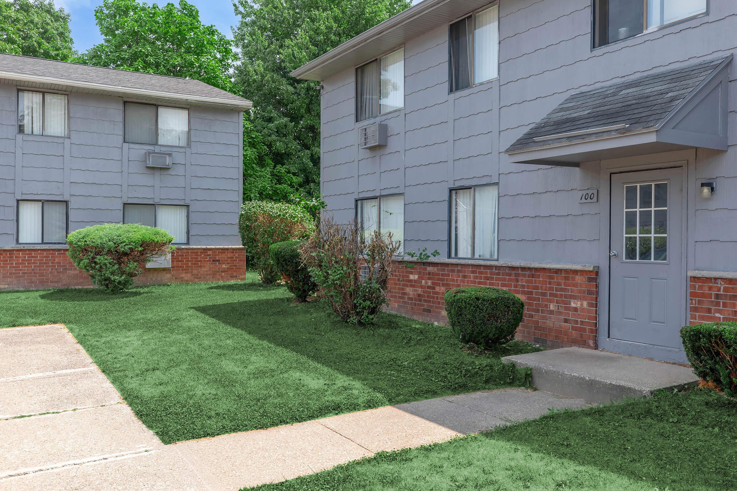 Exterior view of a residential building with two gray multifamily apartments. The foreground features a well-maintained lawn with shrubs and a sidewalk leading to a door marked "104." Background includes another apartment building and greenery in the area.