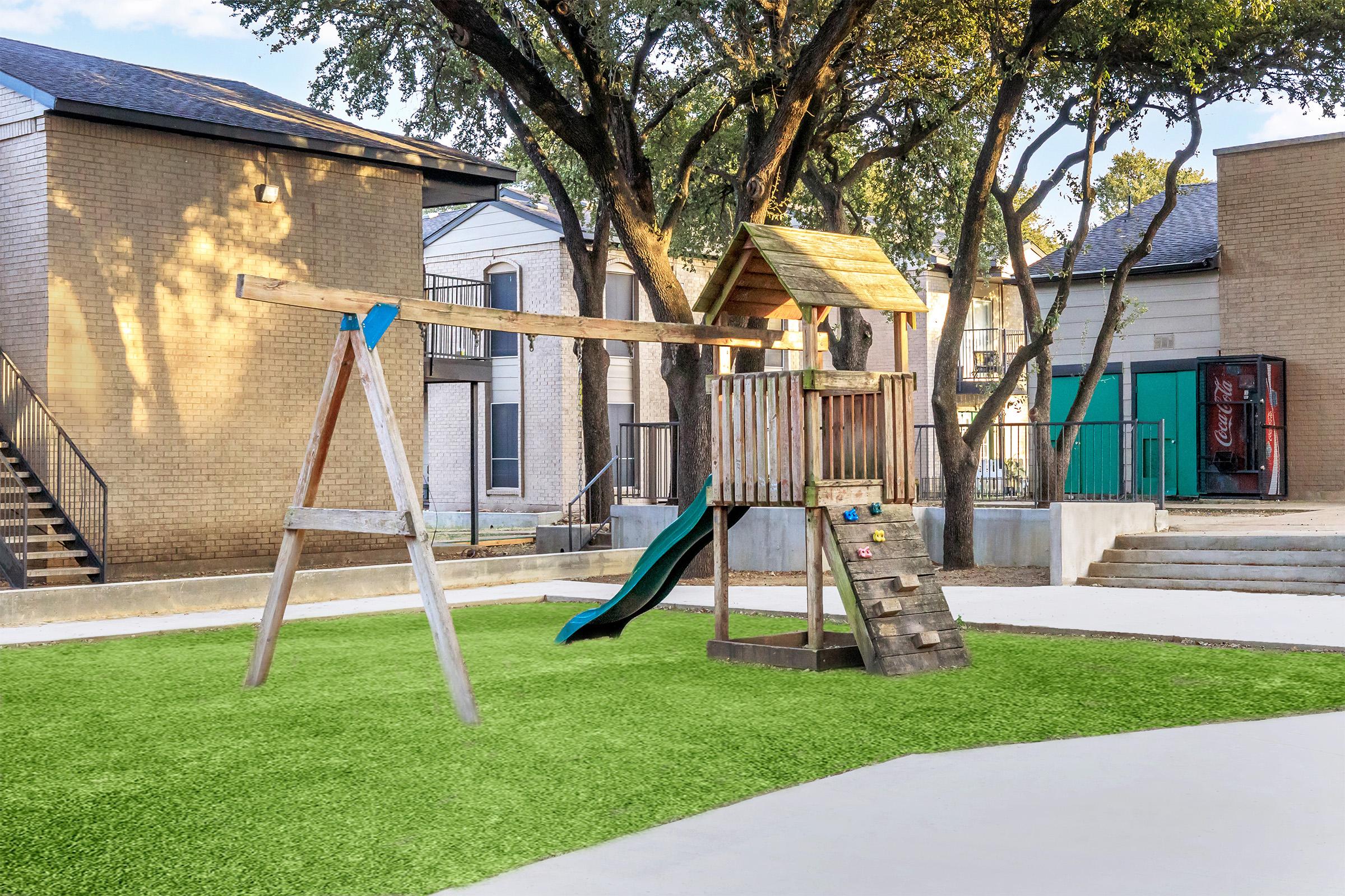 A playground featuring a wooden swing set and a playhouse with a slide, surrounded by green artificial turf. Nearby are trees and buildings, creating a vibrant outdoor space for children to play. A vending machine is visible in the background.