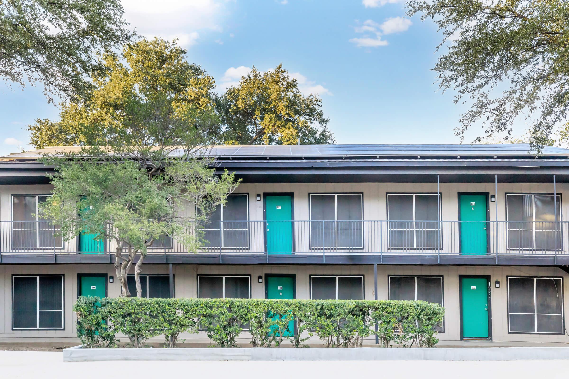 Two-story building with multiple rooms, each featuring a bright turquoise door and large windows. The structure has a modern design with solar panels on the roof. In front, there's a neatly trimmed hedge and trees providing shade under a clear blue sky.
