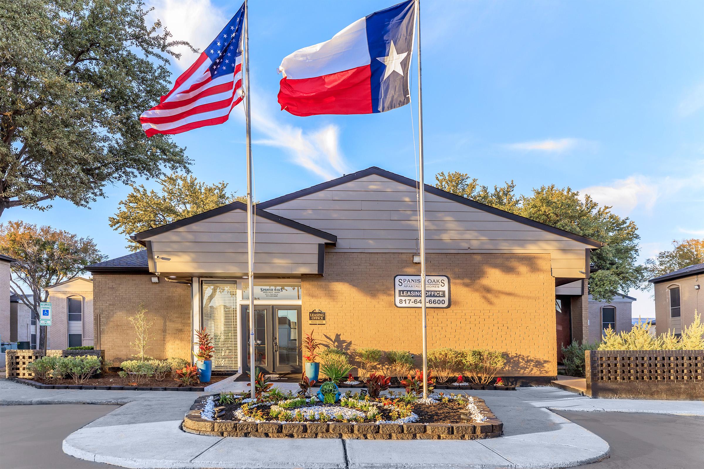 A building entrance with a landscaped garden in front, featuring two flags: the American flag on the left and the Texas flag on the right. The building has a modern design with a welcoming atmosphere, surrounded by trees and a clear blue sky.