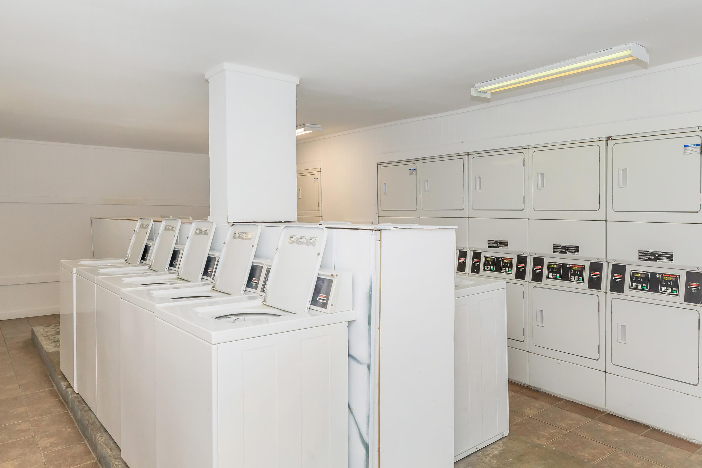 A clean and well-lit laundry facility featuring several white washing machines and stacked dryers against a plain wall. The floor is tiled, and there are overhead lights providing ample brightness.