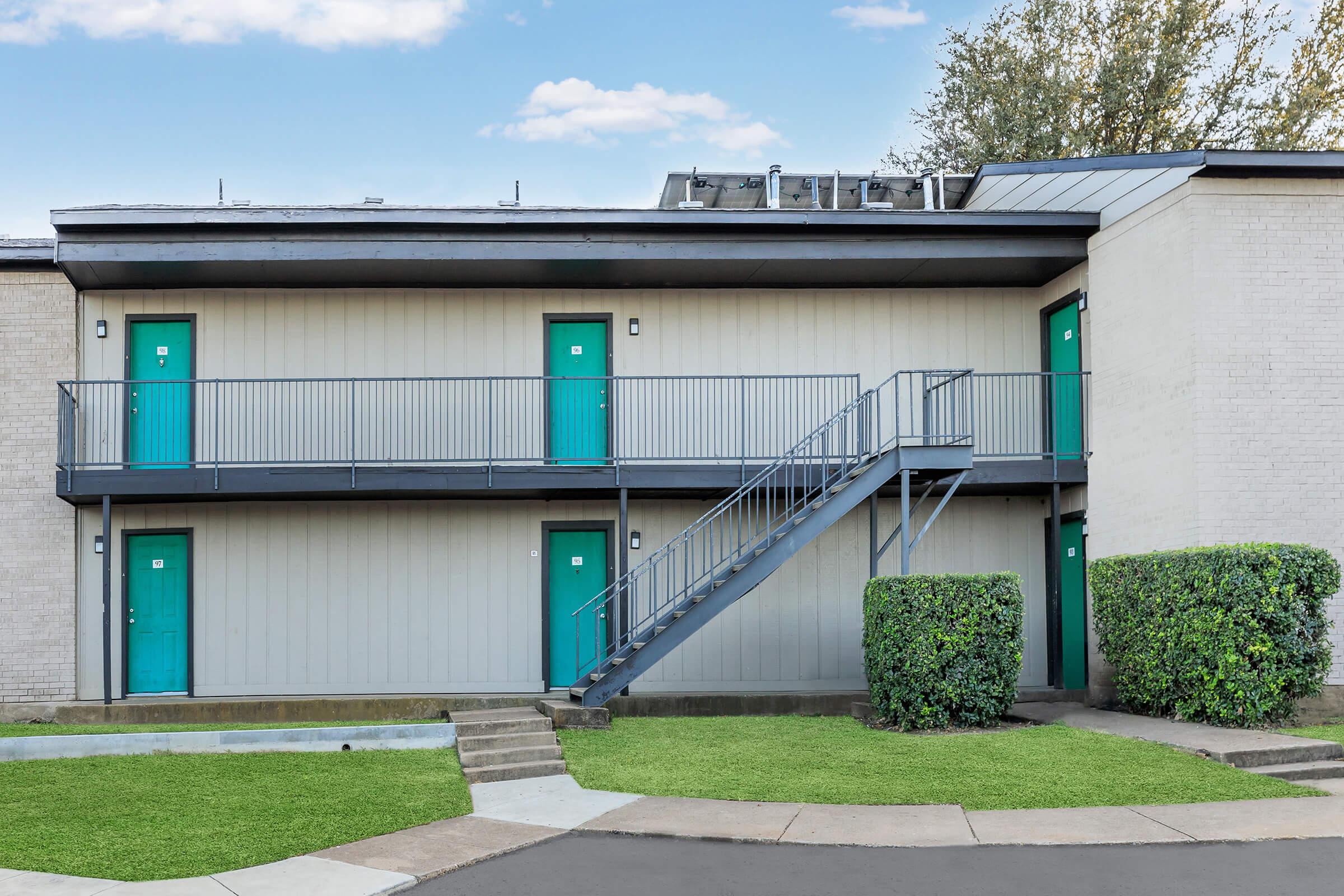 Exterior view of a two-story building with teal doors and a gray metal staircase. Green shrubs flank the entrance, and a small grassy area is visible in front. The sky is clear with light clouds.