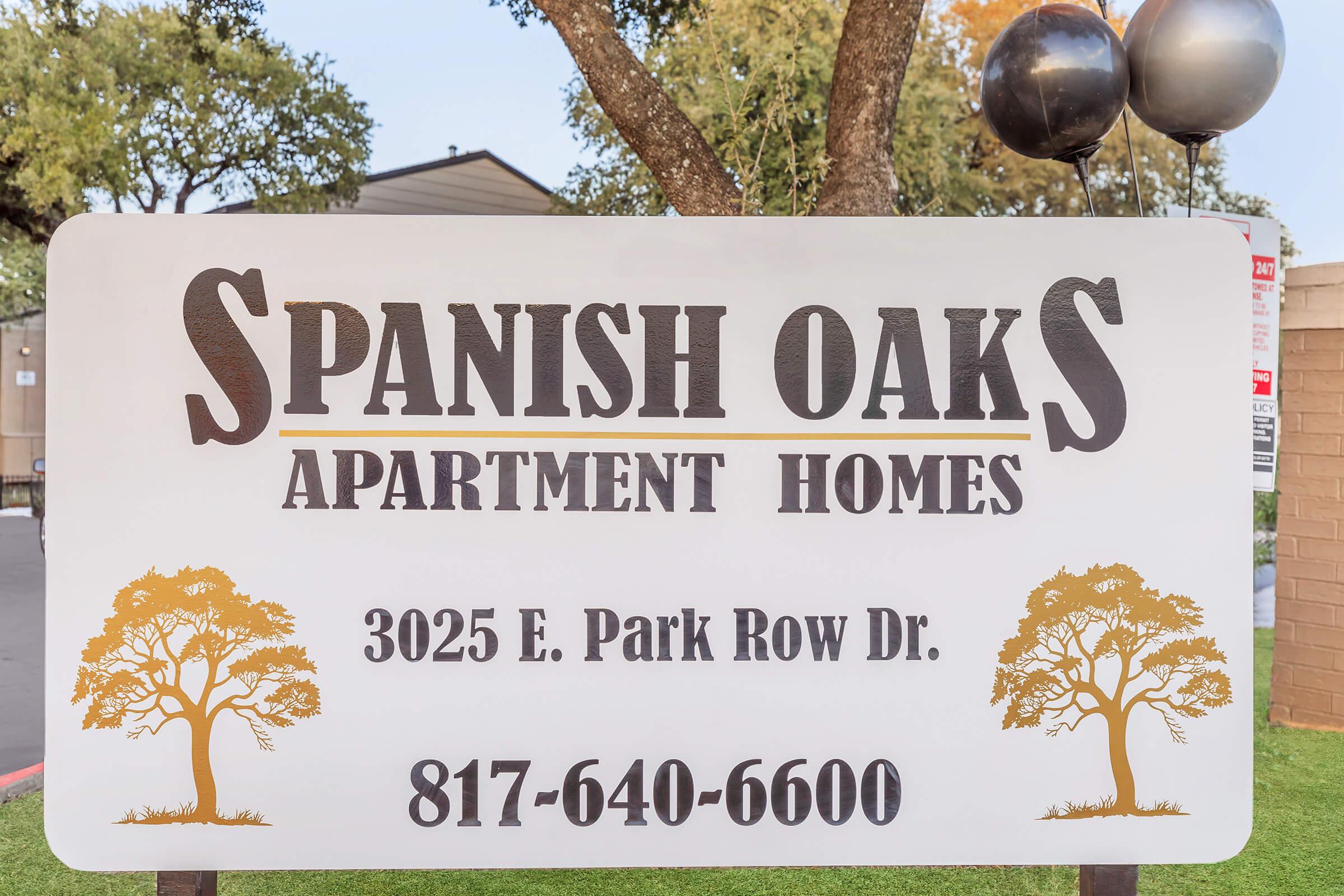 Sign for Spanish Oaks Apartment Homes featuring the address 3025 E. Park Row Dr. and the phone number 817-640-6600. The sign has a decorative design with trees.