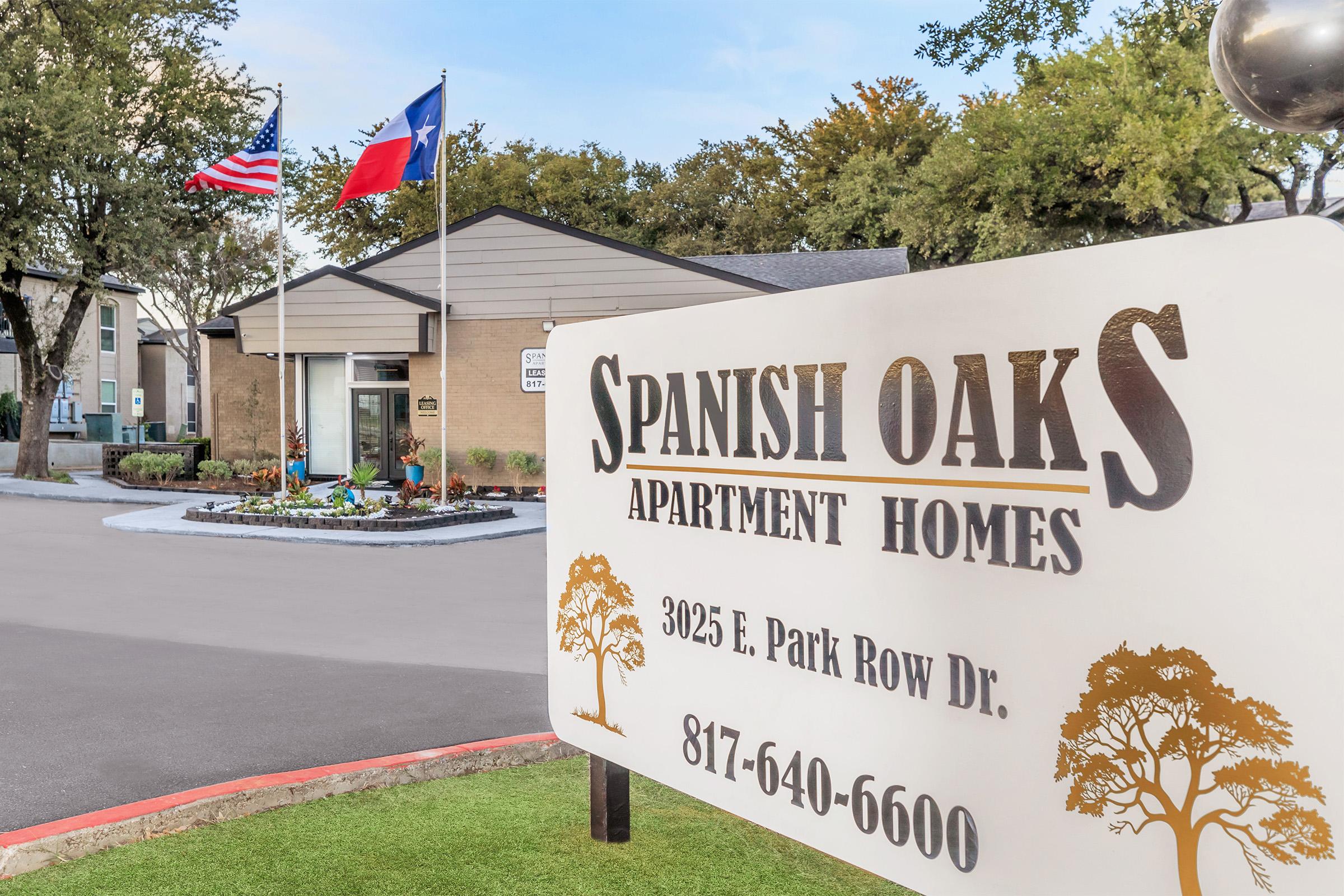 A welcoming view of Spanish Oaks Apartment Homes, featuring a prominent entrance sign, the American and Texas flags flying in front, and well-maintained landscaping. The address is 3025 E. Park Row Dr., with a contact number displayed. The atmosphere is inviting and residential.