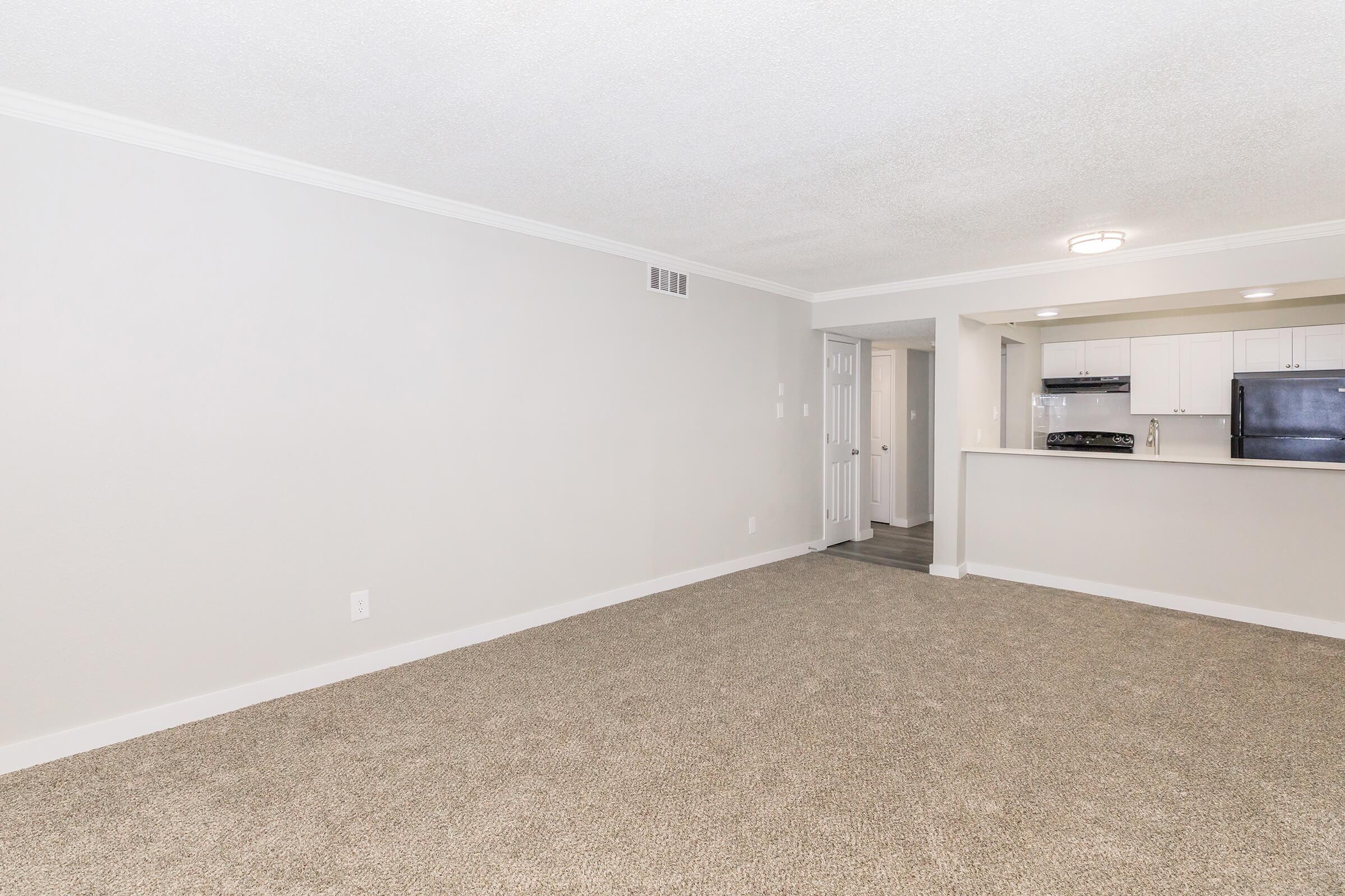 Empty room featuring beige carpet and light gray walls. On one side, there's a kitchen area with modern appliances visible, including a refrigerator and stove. A doorway leads to another room, and the overall space is well-lit with ceiling lights.