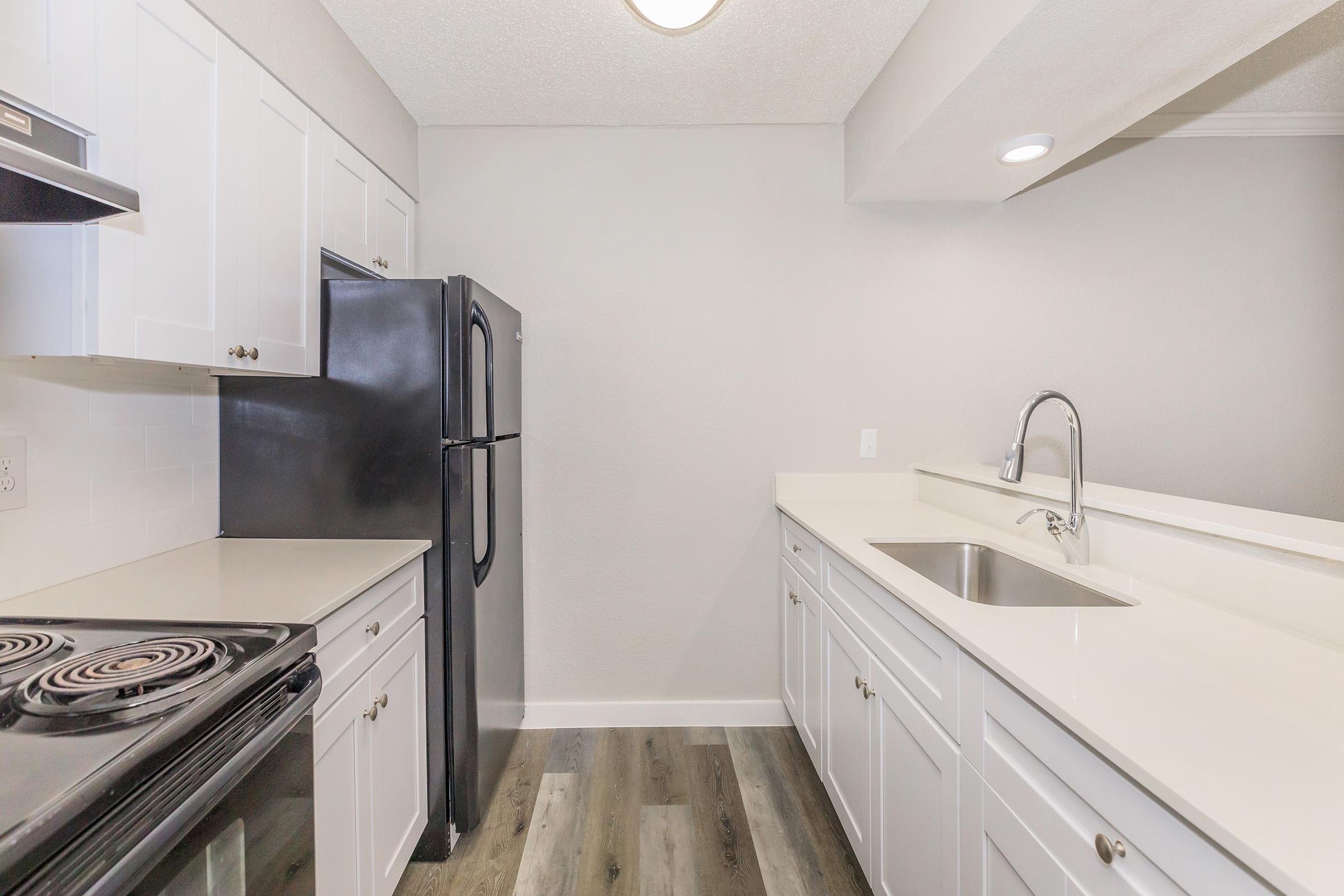 Modern kitchen with white cabinets, a black refrigerator, a black stove, and a stainless steel sink. The countertop is light-colored and the flooring is a wood-like laminate. The walls are painted in a light gray tone, creating a contemporary and clean aesthetic.