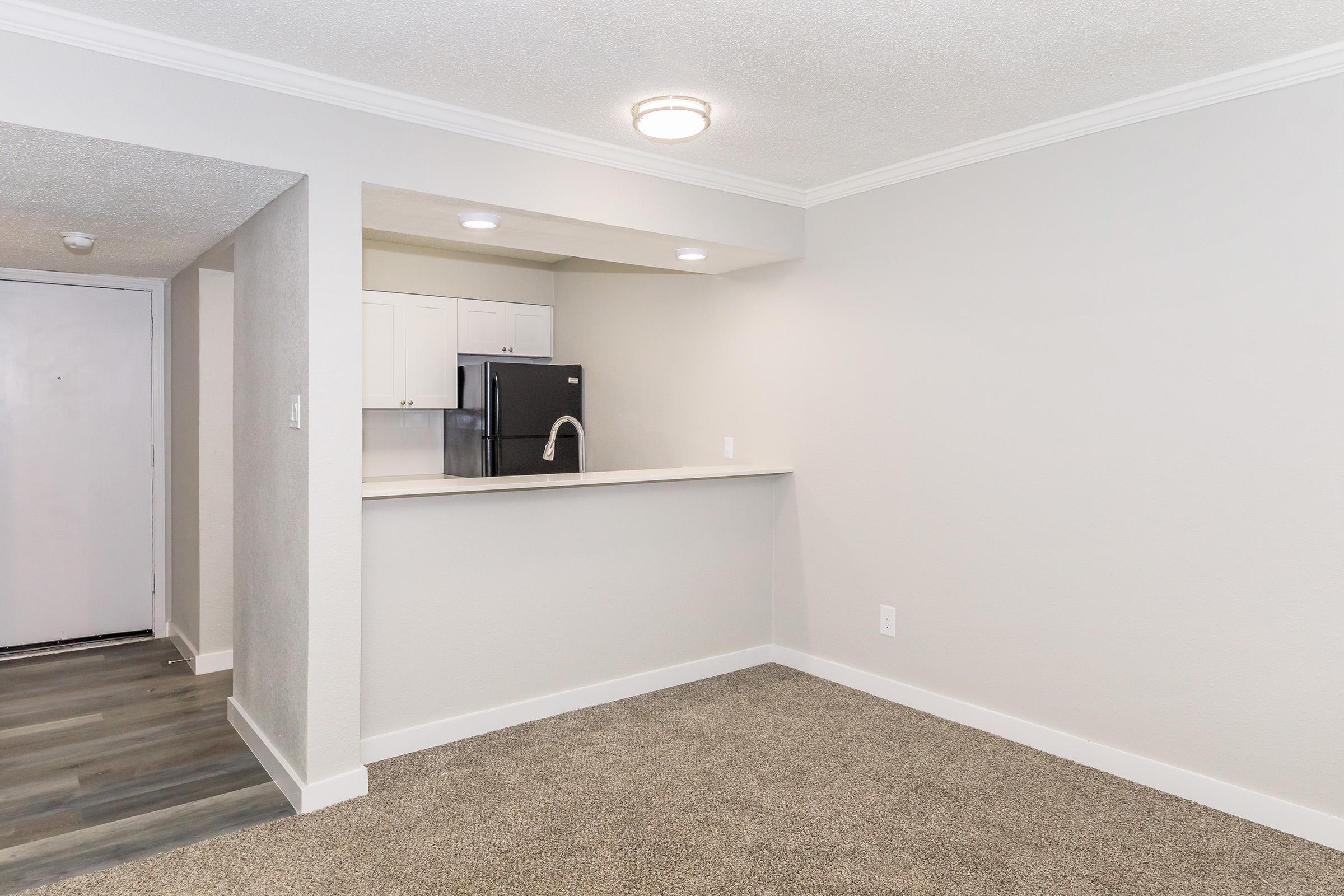 A spacious, well-lit living area featuring gray walls, a plush carpeted floor, and a kitchenette with a black refrigerator. The area has recessed lighting, and a doorway leads to another room. The overall aesthetic is modern and clean, ideal for a cozy apartment setting.