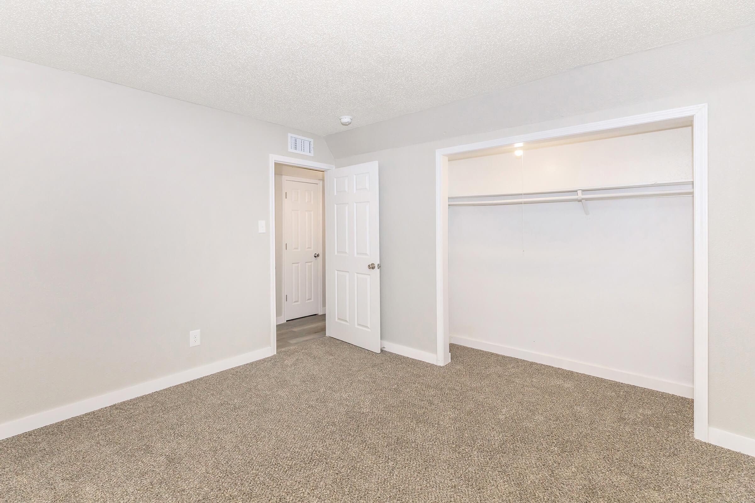 Empty room with light gray walls and a carpeted floor. A closet with a single rod for hanging clothes is visible on the left. A door to another room is seen in the background, providing access to a hallway. The space is well-lit, creating a clean and open atmosphere.