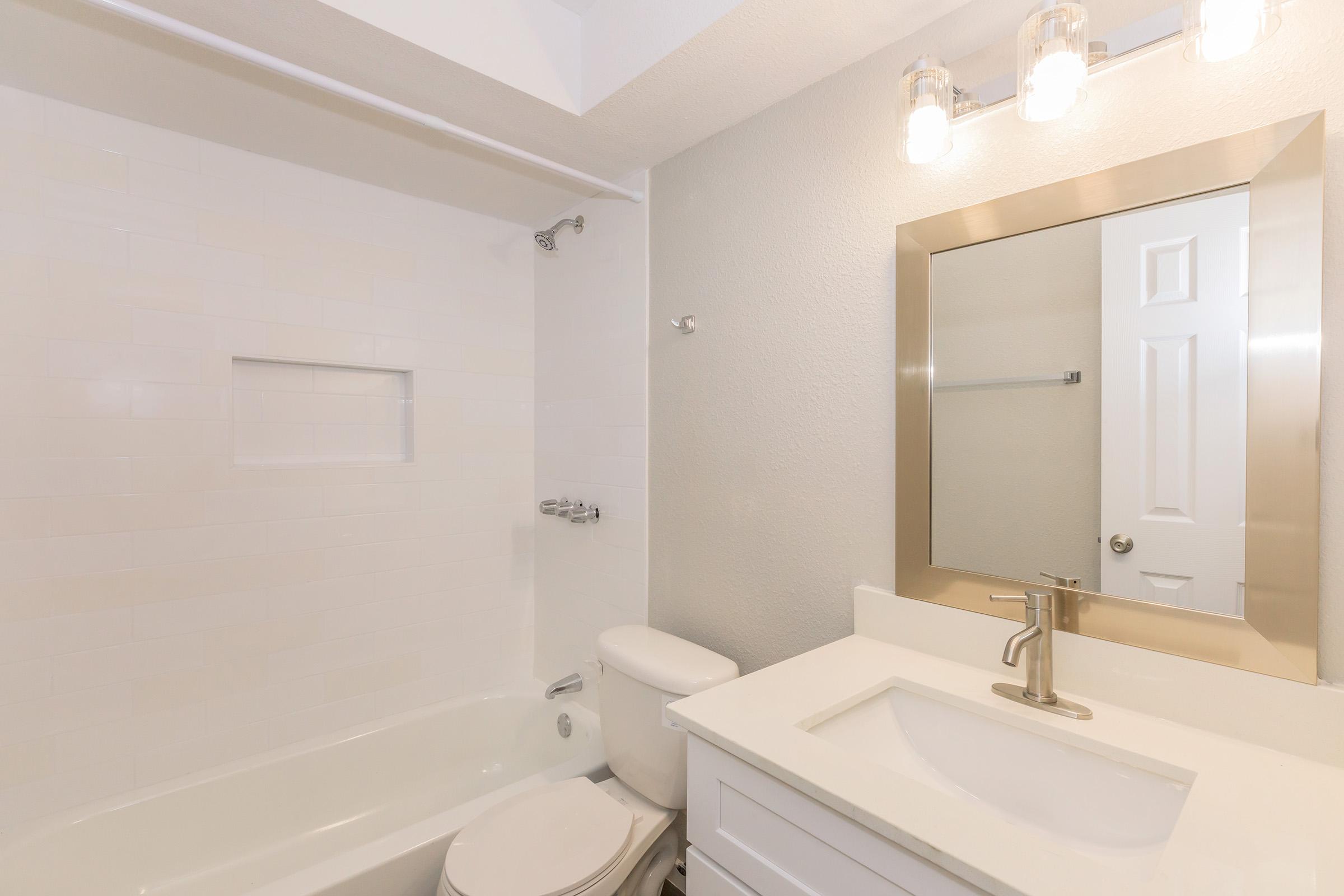 A clean and modern bathroom featuring a white bathtub, a sleek toilet, and a white vanity with a sink. The wall has light-colored tiles and there is a large mirror with a decorative frame. Contemporary lighting fixtures are positioned above the mirror, providing ample brightness in the space.
