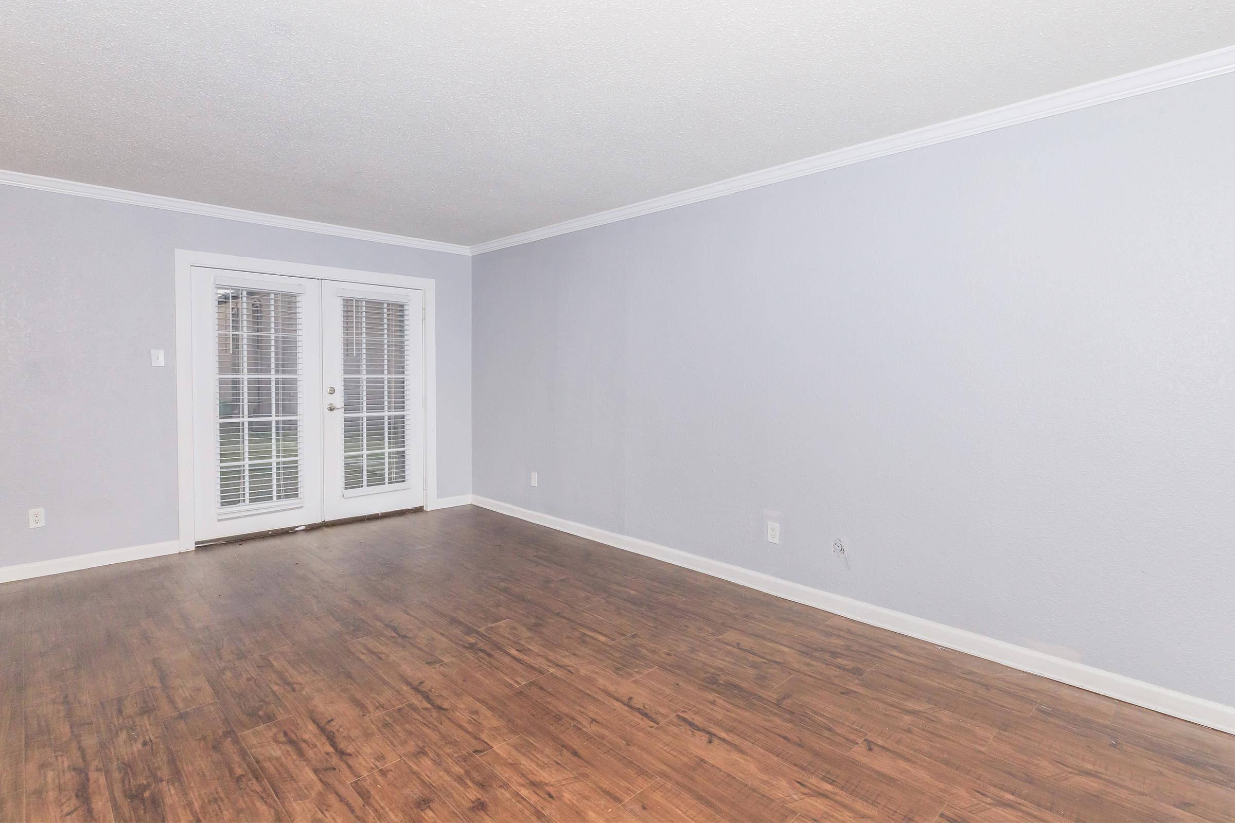 A spacious, empty room with light gray walls and wooden flooring. There is a pair of French doors on one wall, leading outside, and minimal decor. The room appears bright and inviting, with natural light coming through the doors.