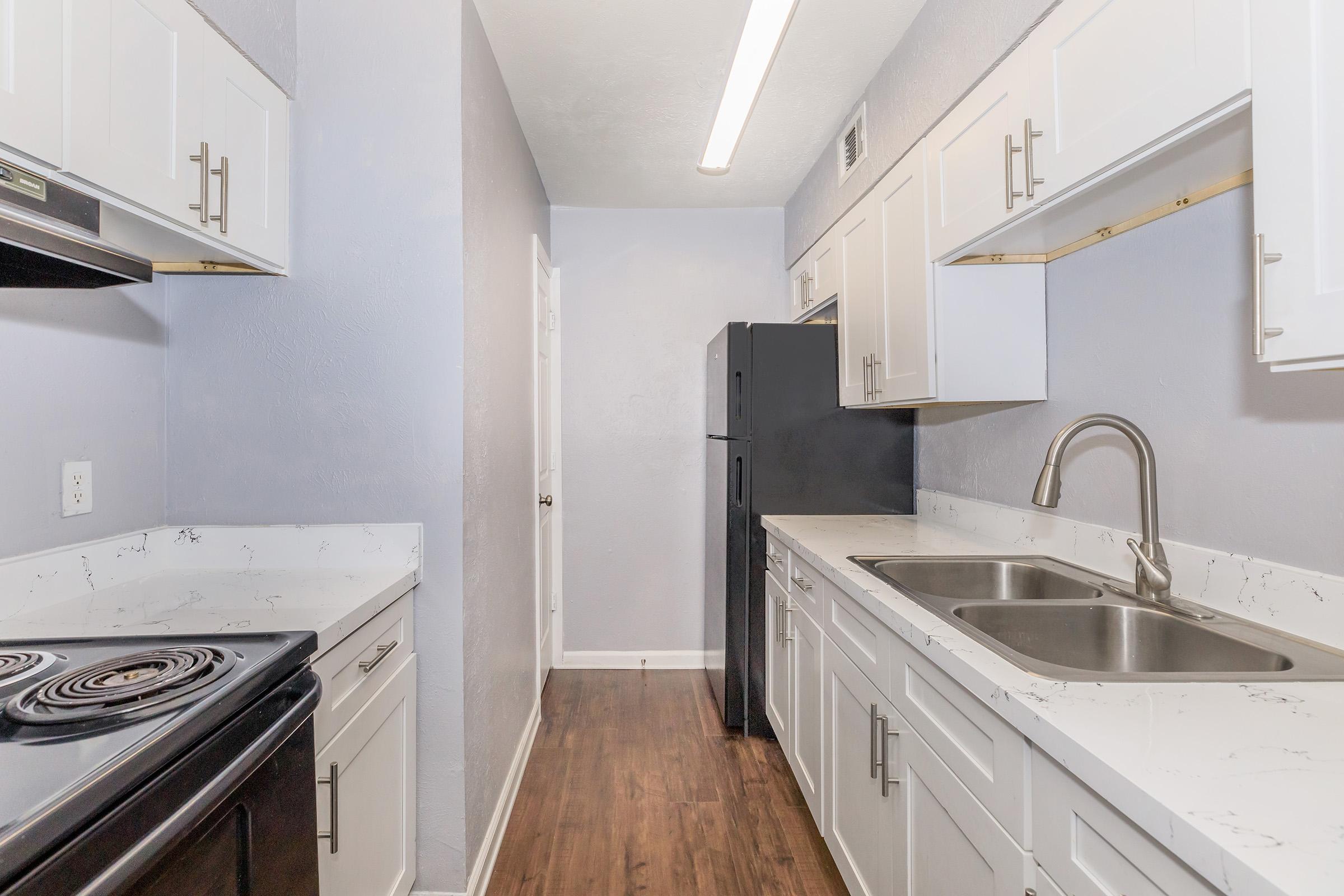 A modern kitchen featuring white cabinets, a black refrigerator, a black stove, and dual stainless steel sinks. The walls are painted light blue, with a smooth countertop and wood-style flooring. A bright overhead light illuminates the space, creating a clean and inviting atmosphere.