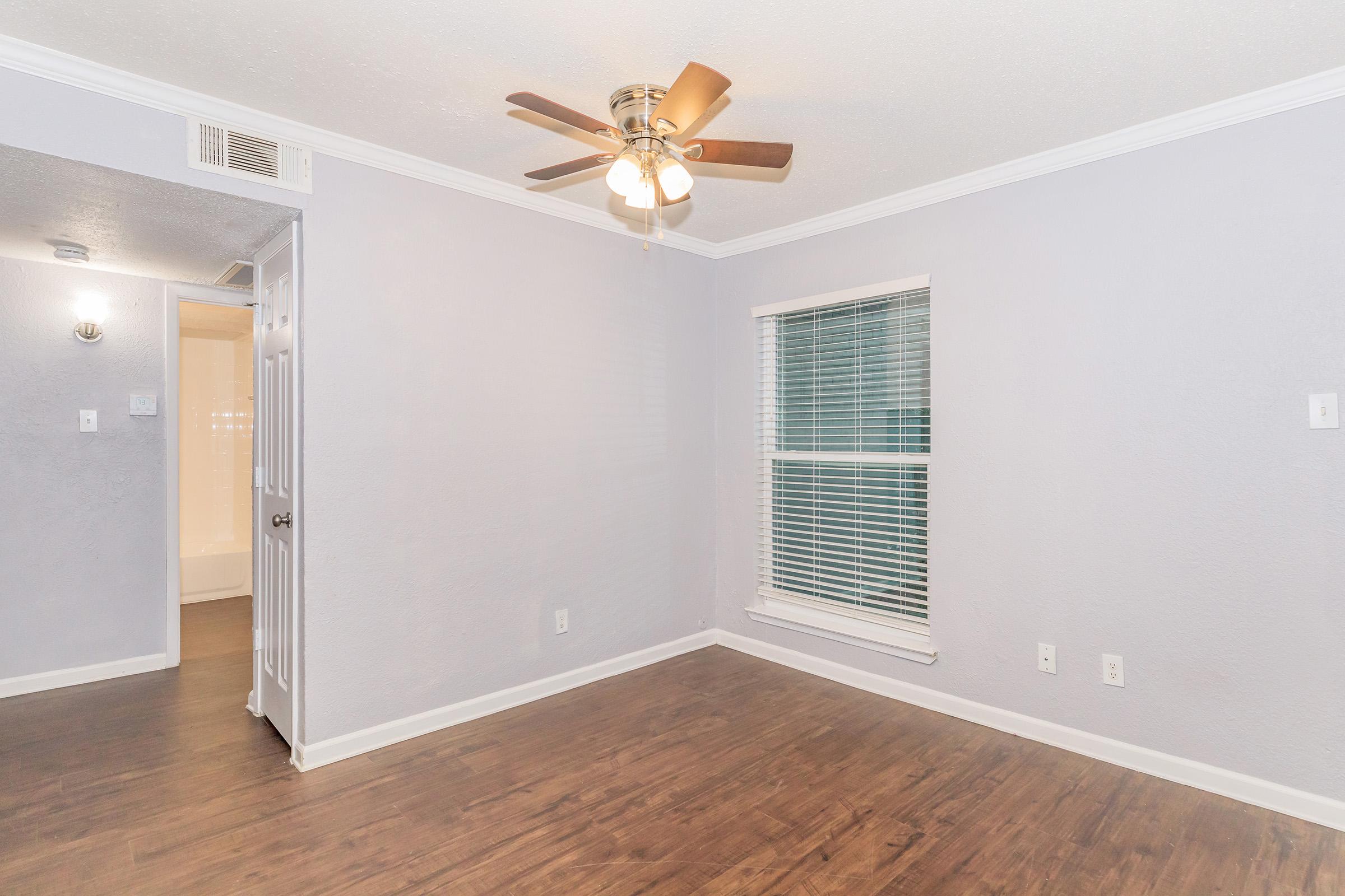 An empty room with a ceiling fan and light fixture, light gray walls, and wooden flooring. A window with white blinds allows natural light, and a door on the left leads to another space. The room has a clean, modern look with a simple design.
