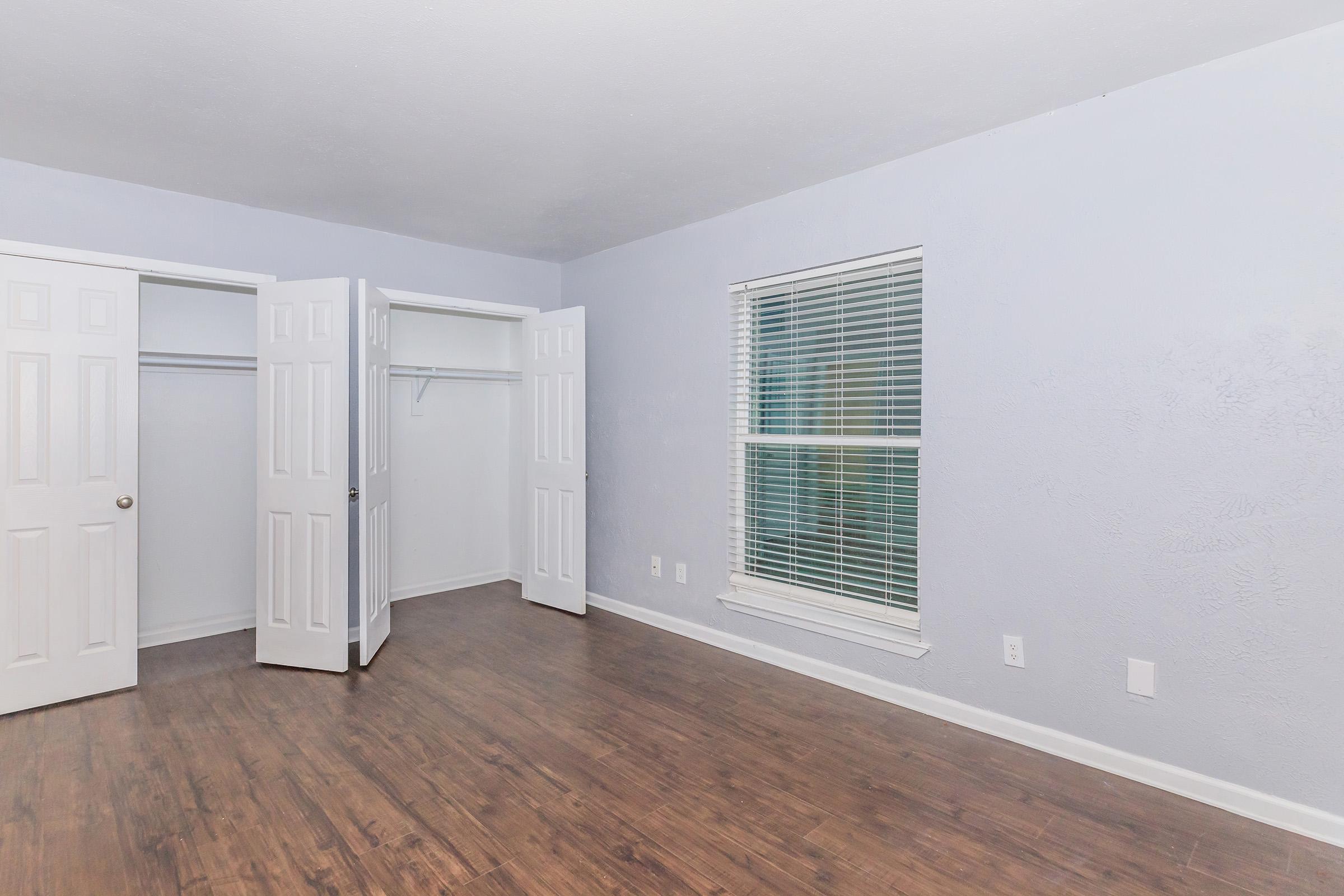 Empty room with light blue walls and wooden flooring. Two open white closet doors on the left side and a window with blinds on the right side. The room has a clean, minimalist appearance, devoid of furniture or decorations.