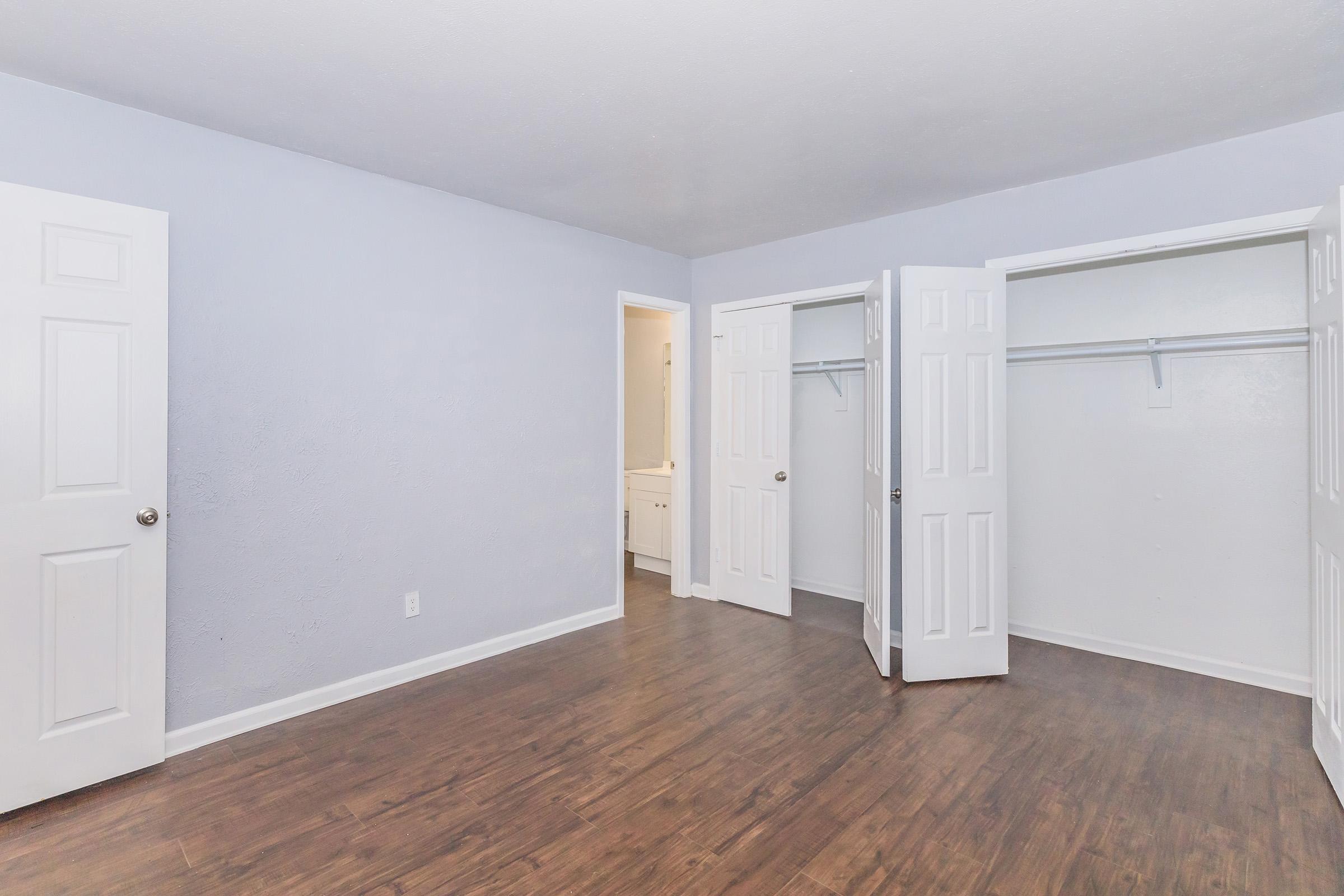 A spacious, empty bedroom featuring light blue walls and a hardwood floor. There are two open closet doors on the right and a doorway leading to a bathroom in the back. The room is illuminated with natural light, providing a clean and inviting atmosphere.