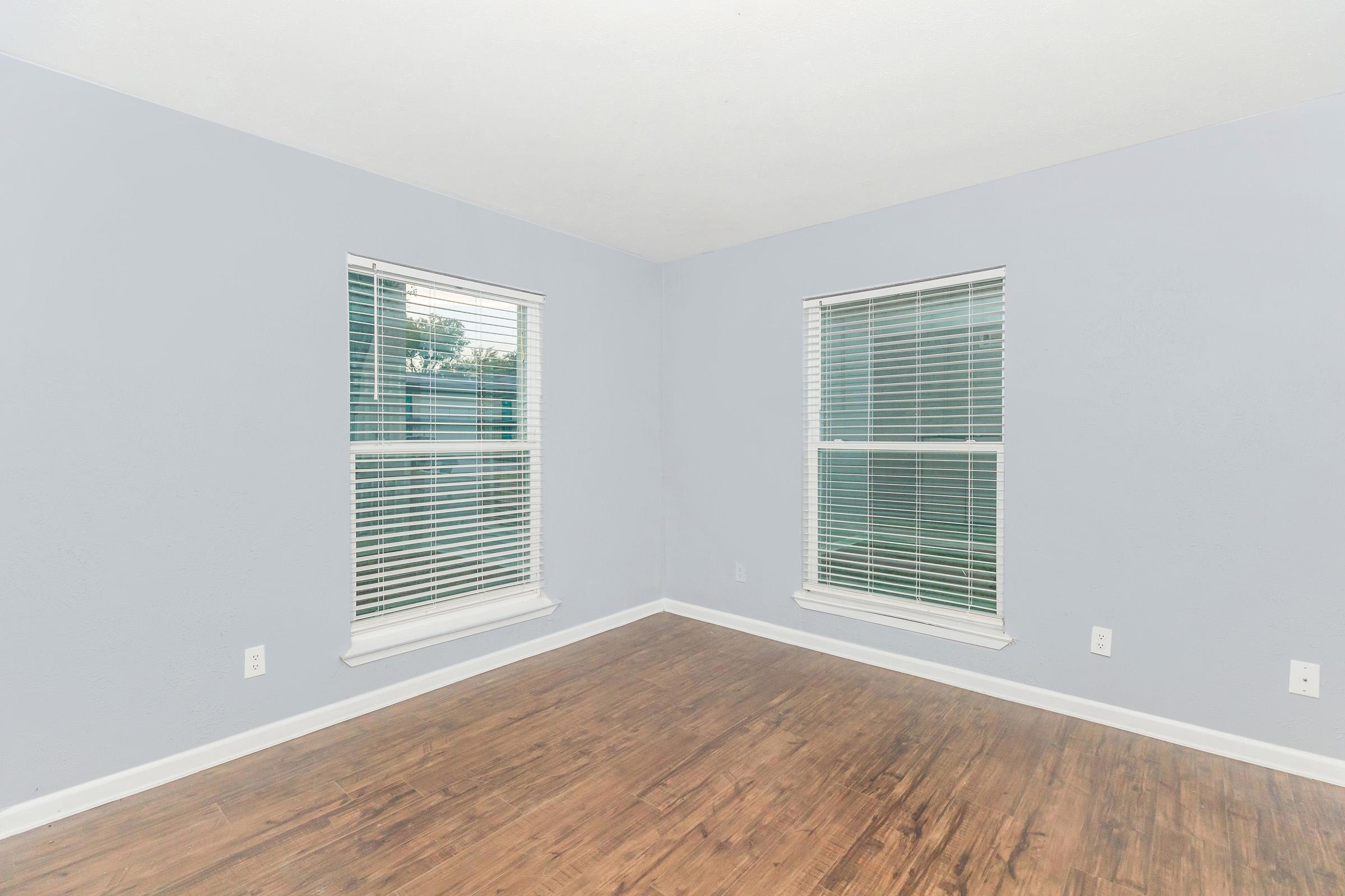 A corner of a vacant room with light blue walls and two windows featuring white blinds. The floor is light brown hardwood, and there are no furnishings or decorations in the space, emphasizing its emptiness and simplicity. Natural light is visible coming through the windows.