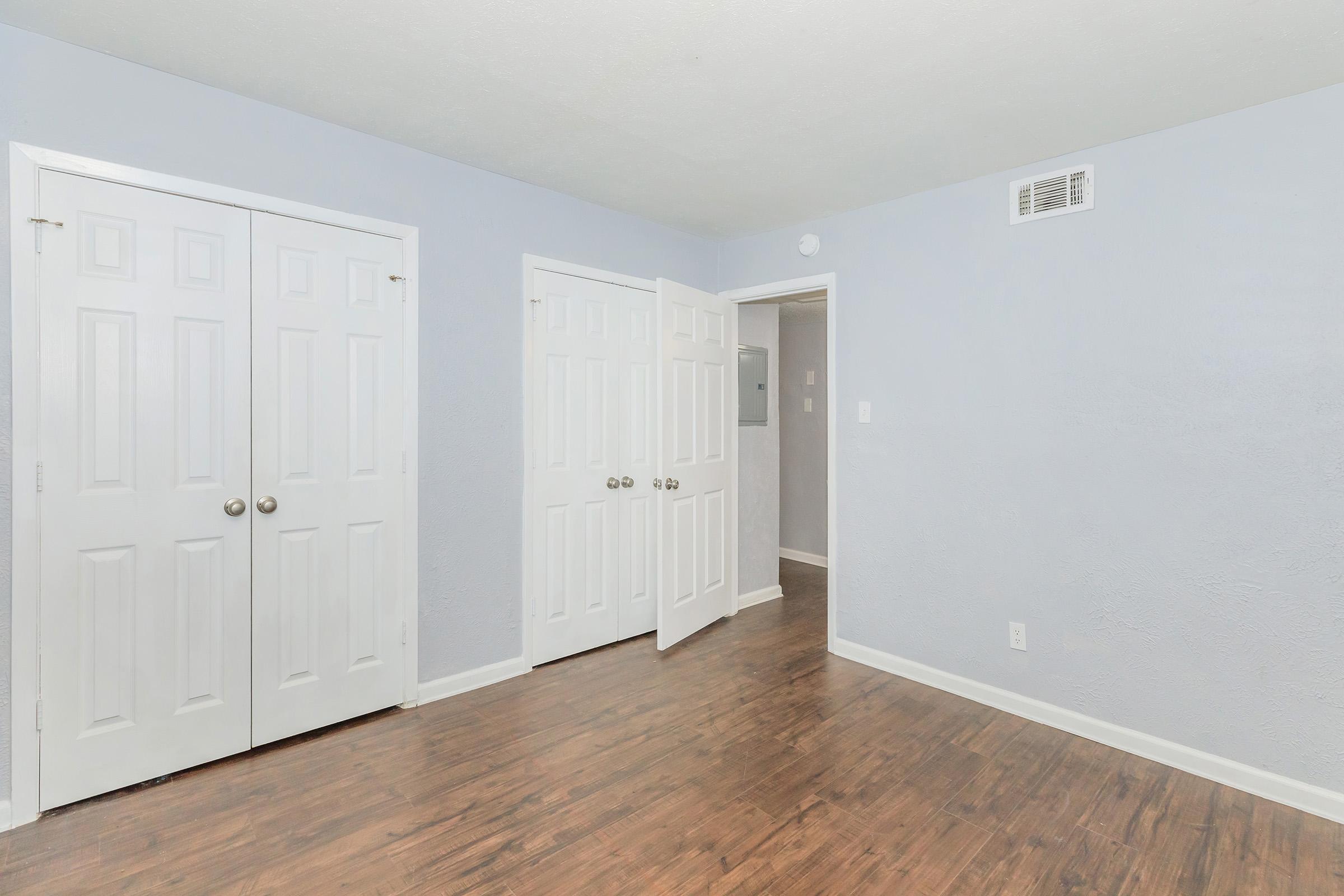 An interior view of a light blue room featuring two white doors with round knobs, a closet on the left, and a doorway on the right leading to another space. The floor is a warm wood tone, and the walls are painted in a soft, neutral color. The room is well-lit and has a clean, tidy appearance.