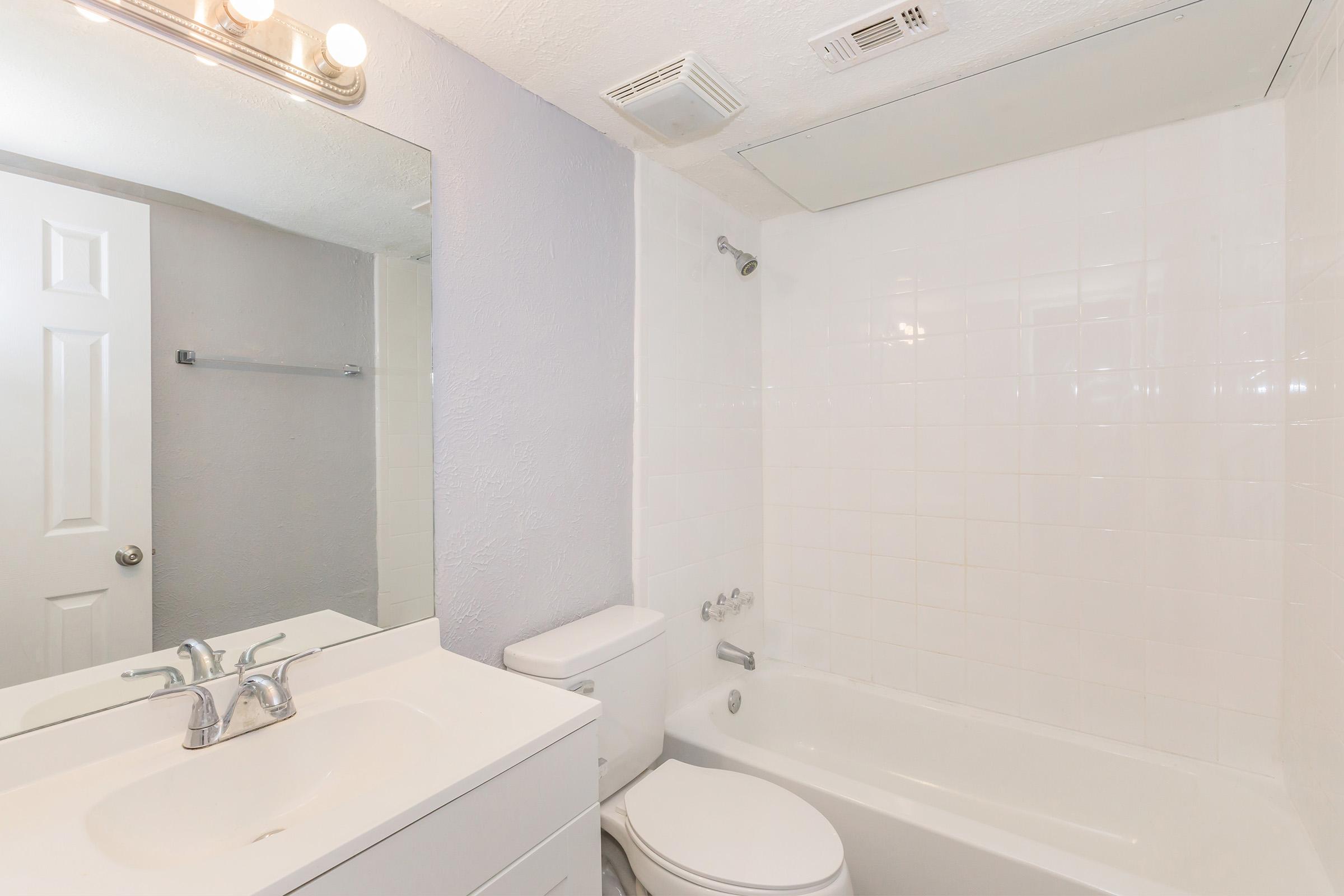 A clean, modern bathroom featuring a white tiled bathtub, a toilet, and a sink with a sleek faucet. The walls are painted in a light color, and there is a large mirror above the sink. A light fixture with multiple bulbs is mounted above the mirror, providing ample lighting. A towel rack is visible on the wall.