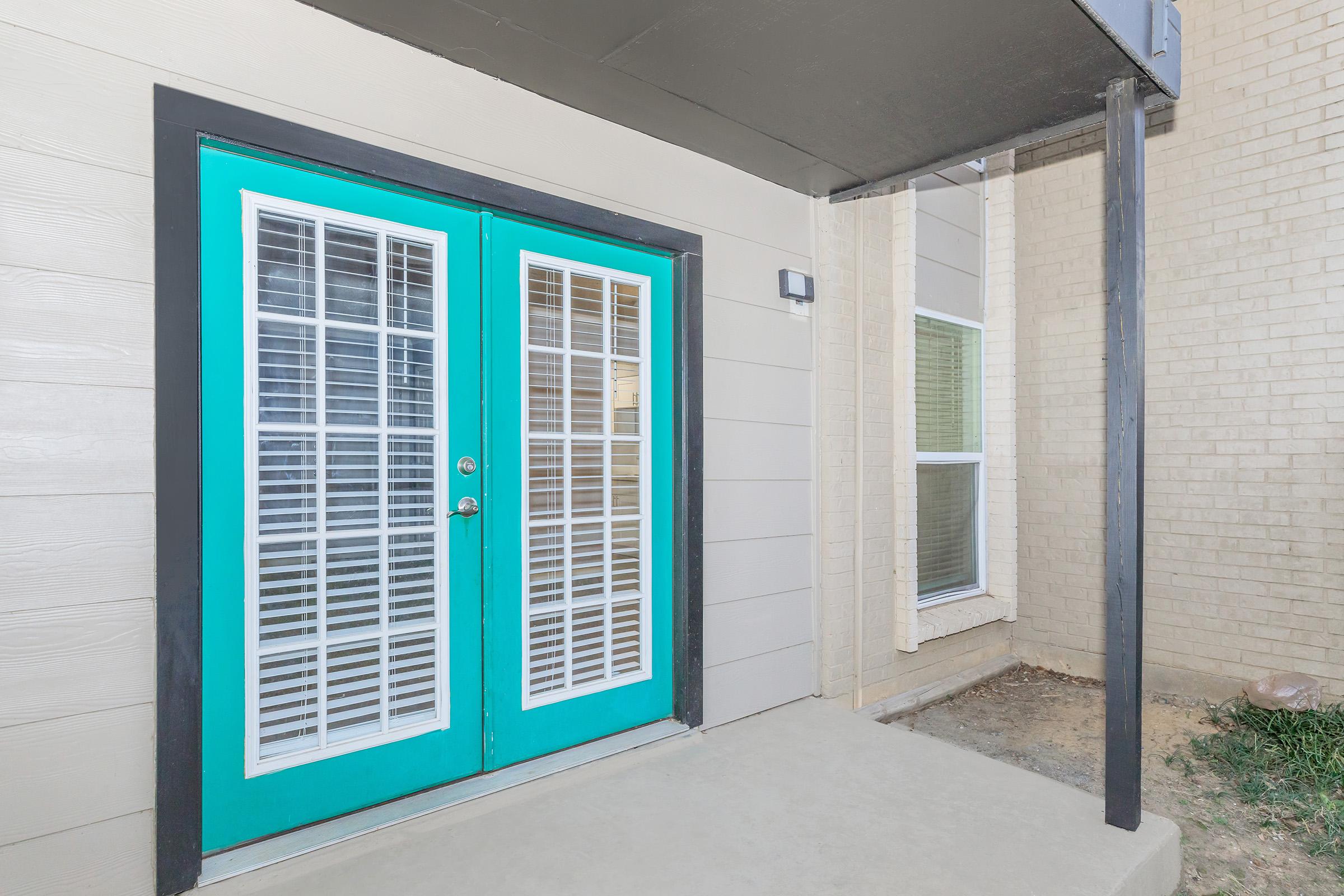 A pair of bright turquoise double doors with white shutters, framed by a black border, situated at the entrance of a building. The doors are set against a light beige exterior wall, with a small porch area and a vertical black support post on the side. Windows are visible to the right.