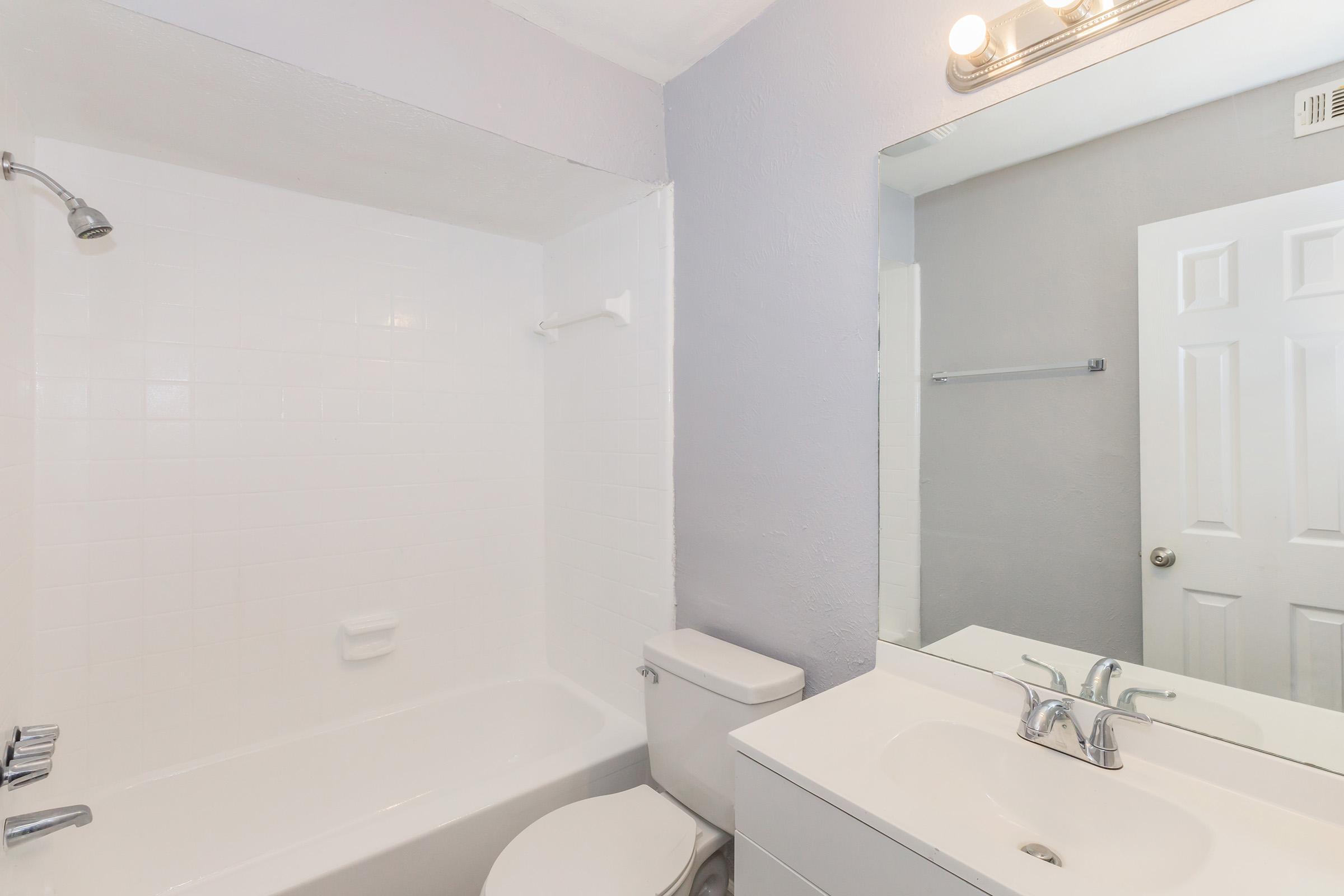 A clean, well-lit bathroom featuring a white bathtub with a showerhead, a toilet, and a modern sink with a mirror above. The walls are painted in light gray, and there is a door leading to another room. Overall, the space is neatly organized and inviting.