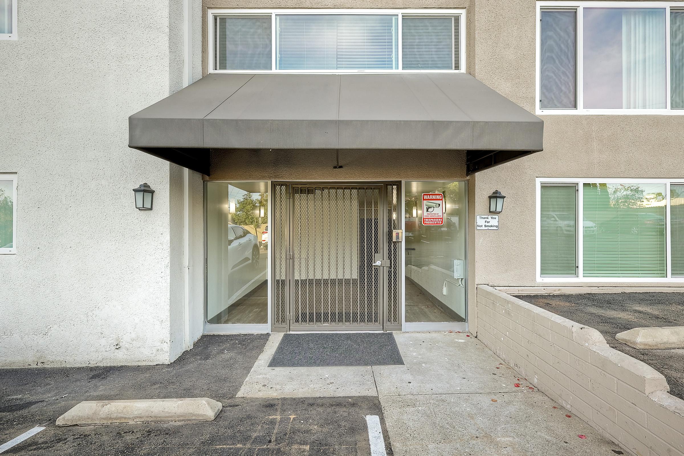 Entrance of a building with a glass door and a gray awning. The entrance is secured with a metal gate. Windows on either side are covered with blinds. A "No Trespassing" sign is visible near the door, along with light fixtures on the wall. The pathway is concrete, with leaves scattered on the ground.