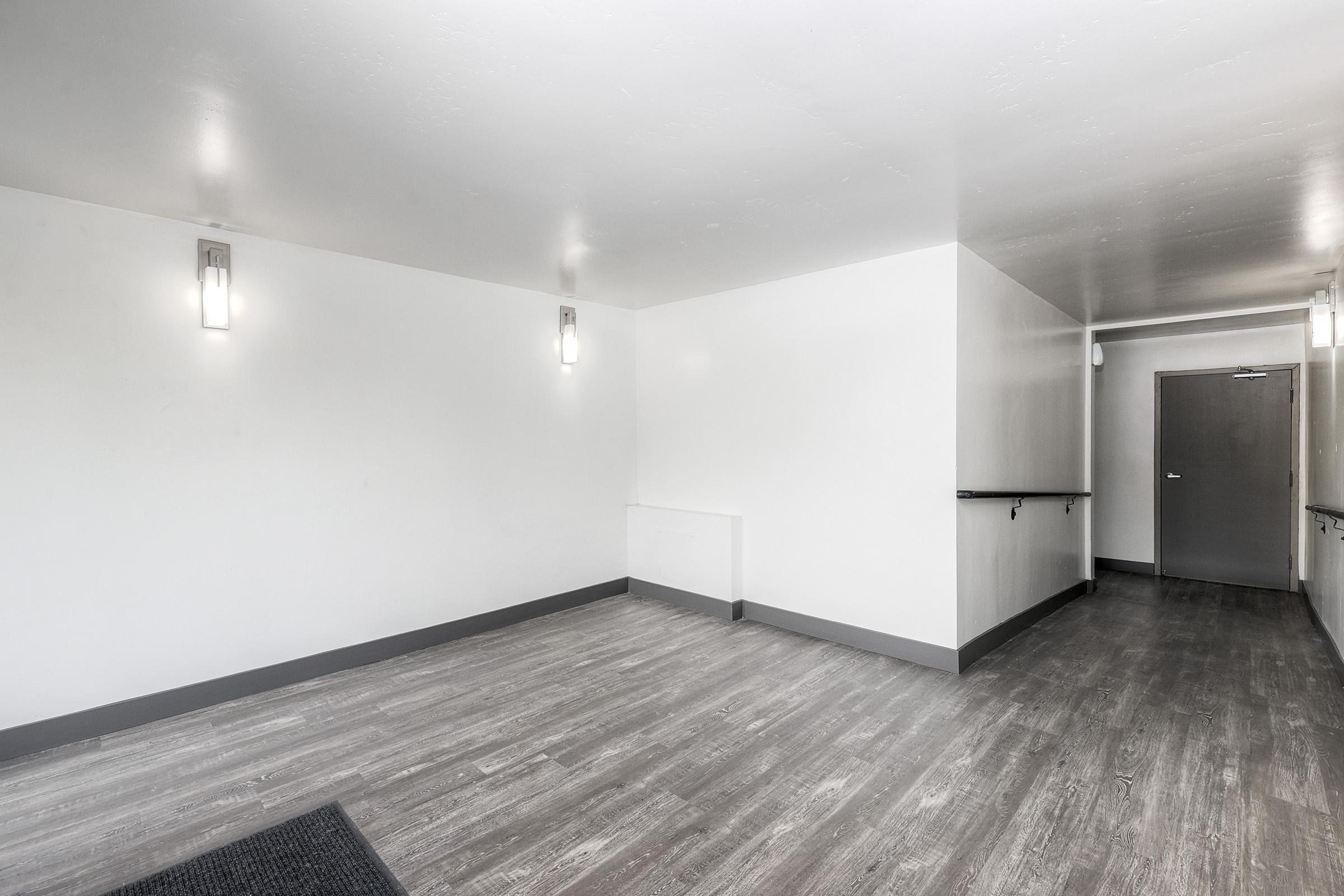 Empty interior space with white walls, recessed lighting, and a smooth gray floor. There is a doorway on the right side leading to another room, and a short wall with a railing on the left. The overall atmosphere is bright and minimalistic.
