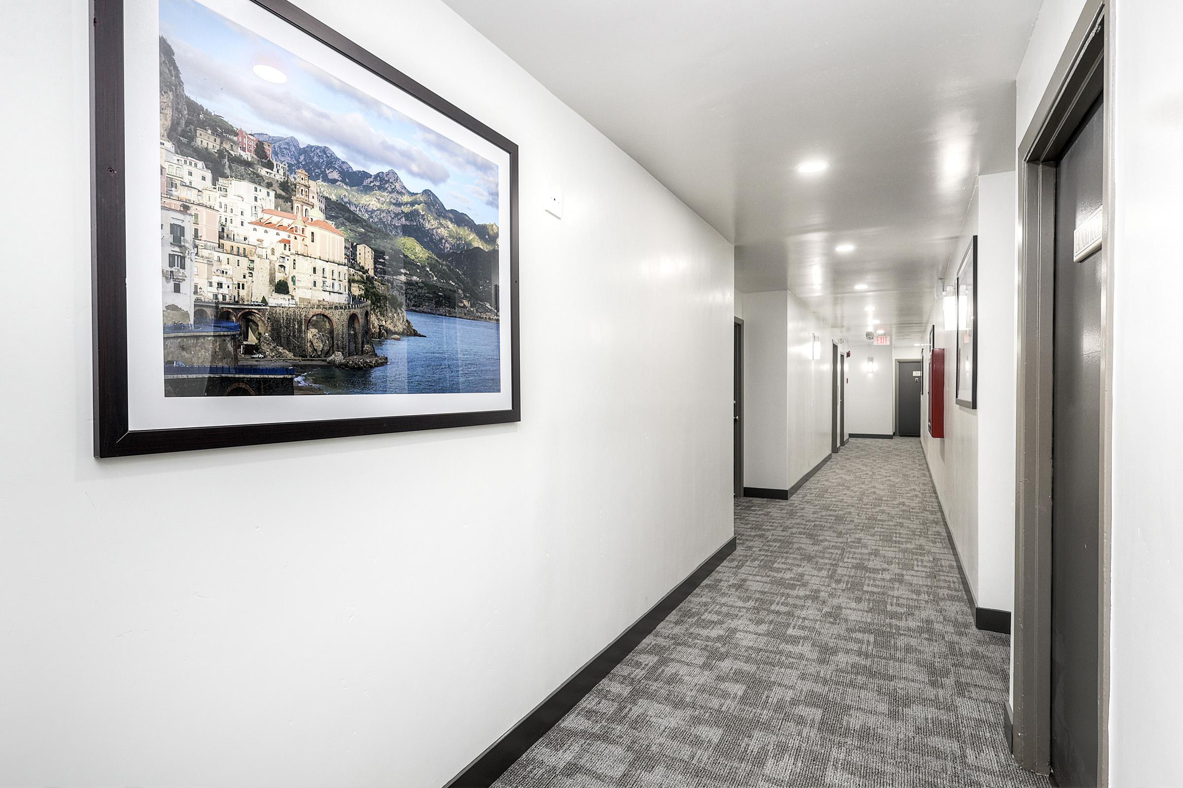 A well-lit corridor featuring light gray walls and a patterned carpet. On one wall, there is a large framed photograph of a coastal town with mountains in the background. The opposite wall has multiple doors, leading to different rooms, contributing to a clean and modern interior design.