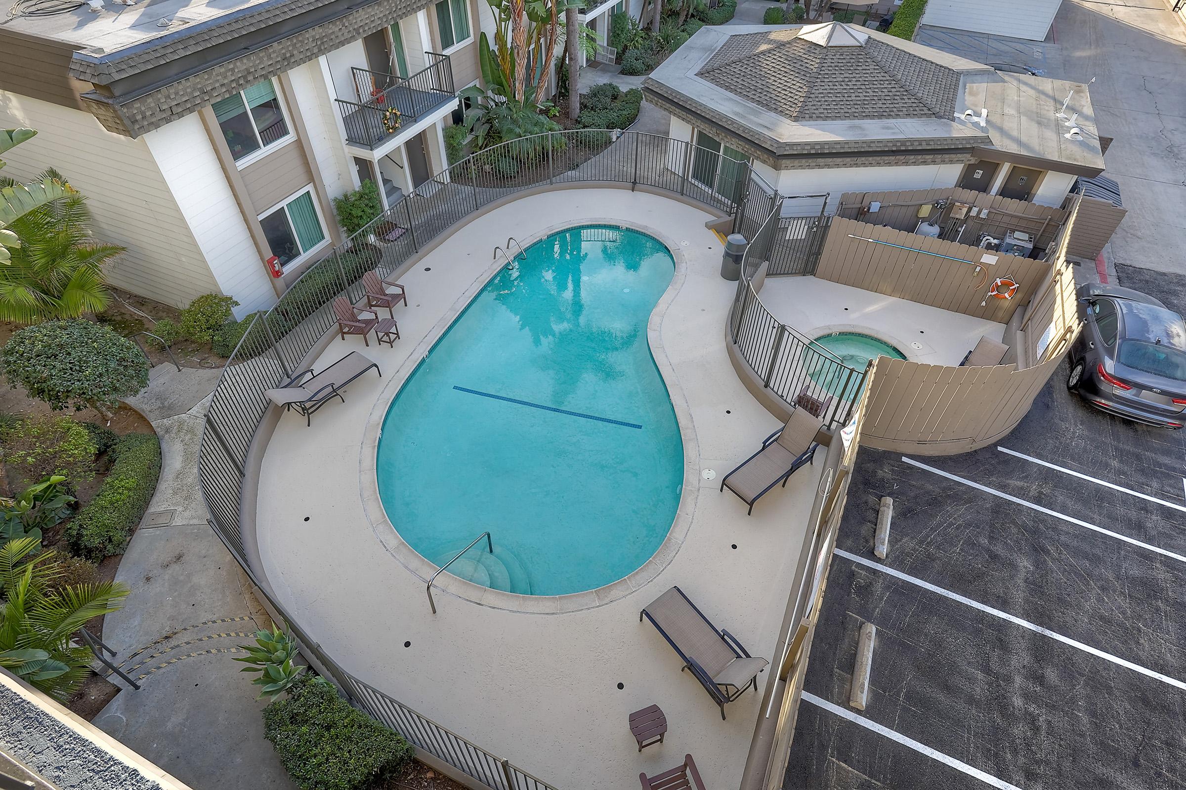 Aerial view of a swimming pool area featuring an irregularly shaped pool with lounge chairs around it, surrounded by a fence. A hot tub is adjacent, and several apartment buildings are visible in the background. There is a parking area with cars nearby, and greenery enhances the setting.