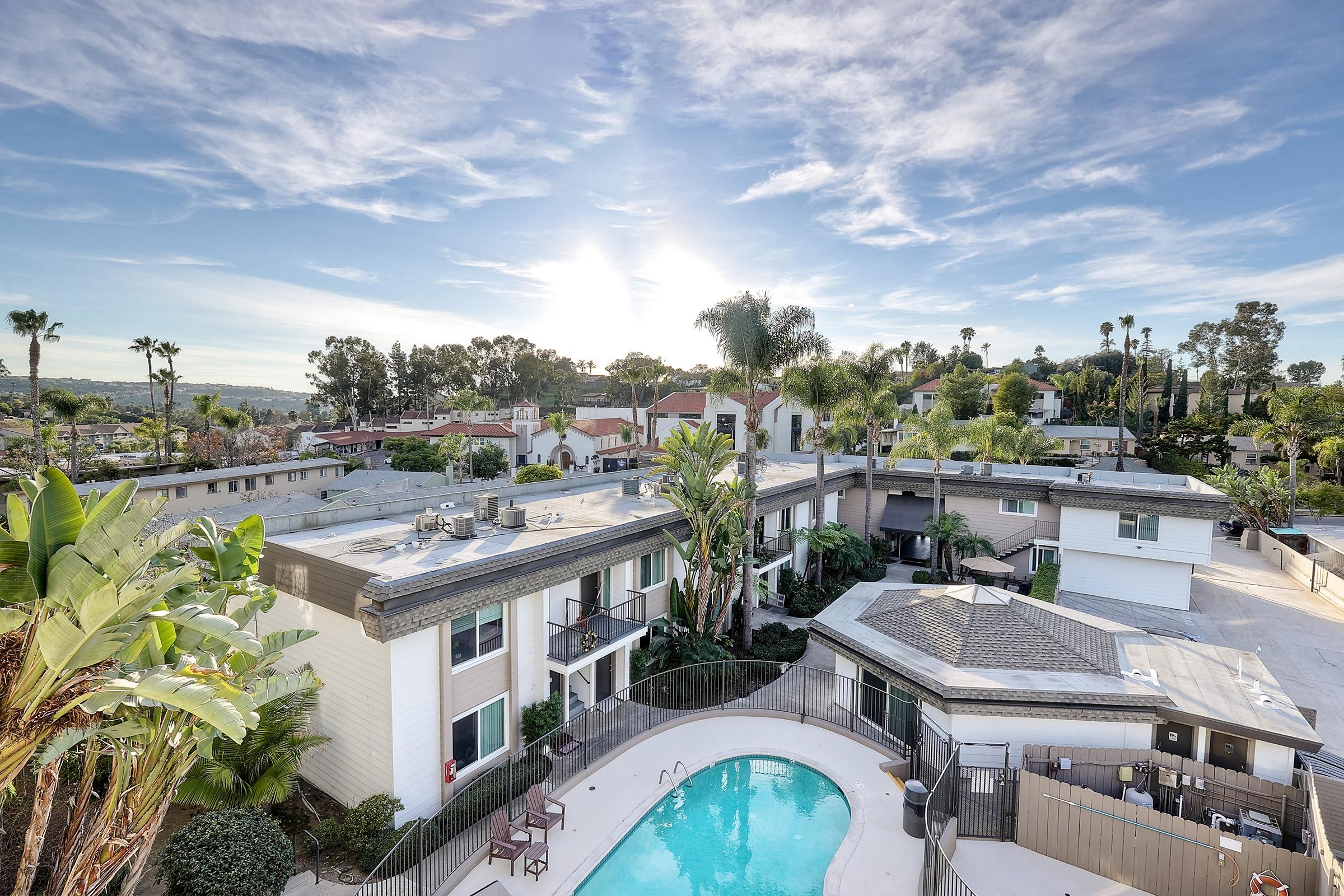 Aerial view of a residential complex featuring a swimming pool surrounded by palm trees. The sun sets in the background, creating a warm glow over the buildings and landscaped area. The scene conveys a relaxed, tropical atmosphere with clear skies and greenery.