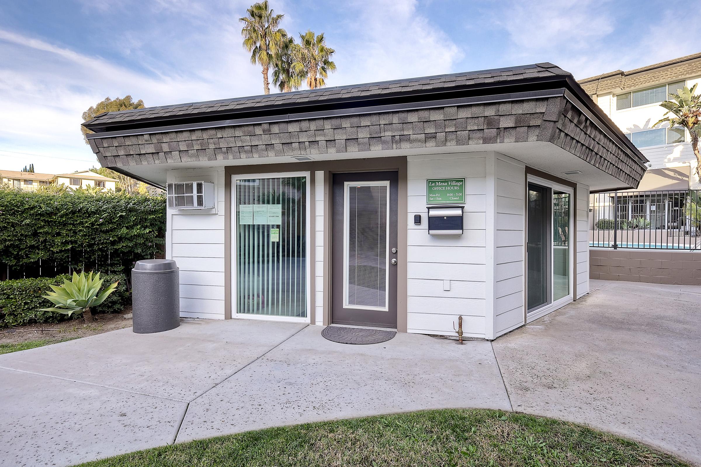 A small, modern building with a flat roof, featuring glass windows and a door. It has a sign near the entrance, a mailbox on the side, and landscaping with palm trees and plants. The building is situated in a well-maintained outdoor area, with a glimpse of a swimming pool visible in the background.