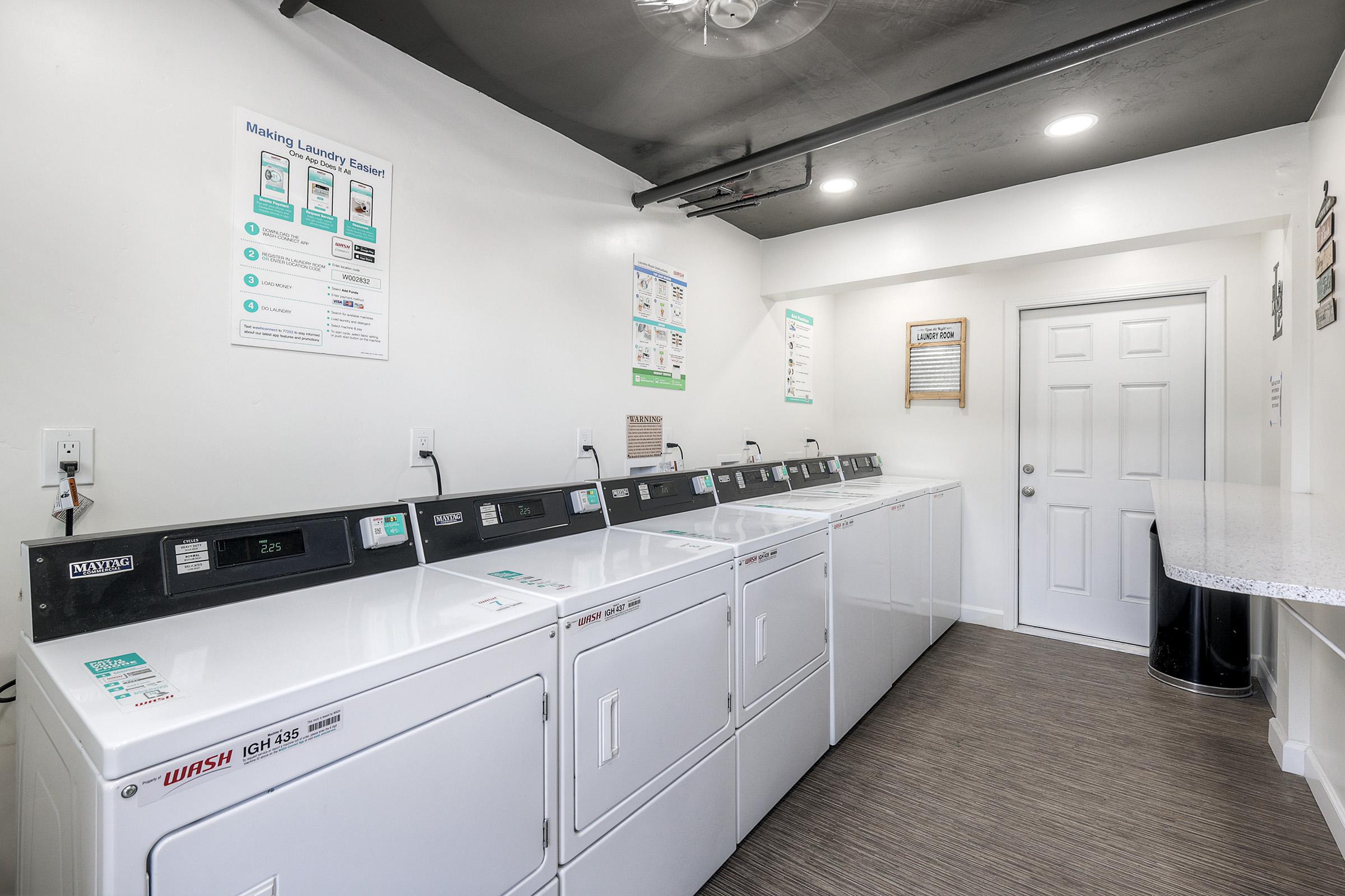 A clean and modern laundry room featuring multiple white washing machines and dryers lined up against a wall. The room has bright lighting, a poster with laundry instructions on the wall, and a door leading to another area. A dark trash bin is also visible in the corner.