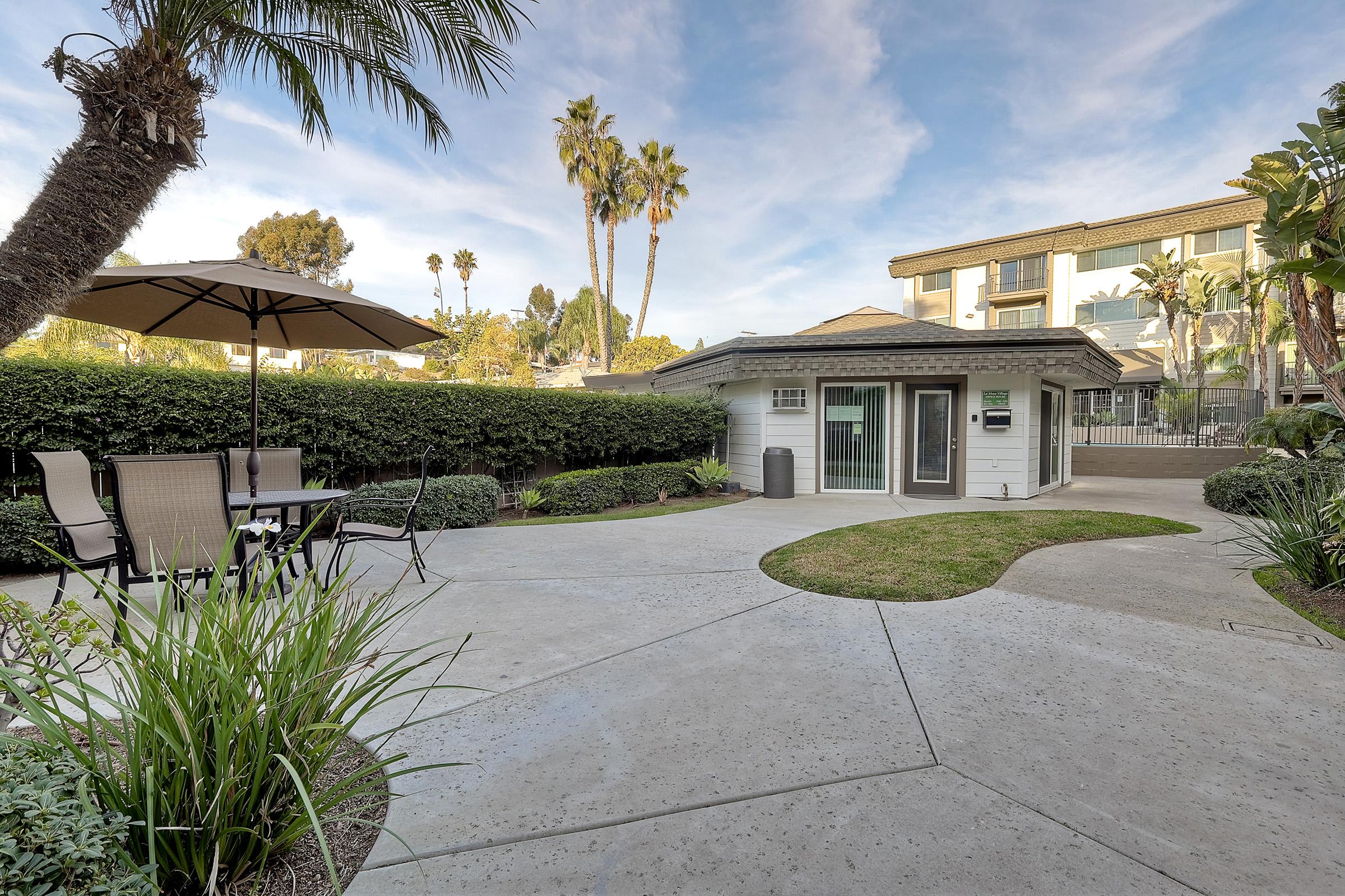 A serene outdoor space featuring a patio with a table and chairs under an umbrella, surrounded by lush greenery and palm trees. In the background, a modern building is visible, adding to the tranquil atmosphere. The sky is partly cloudy, enhancing the inviting ambiance.