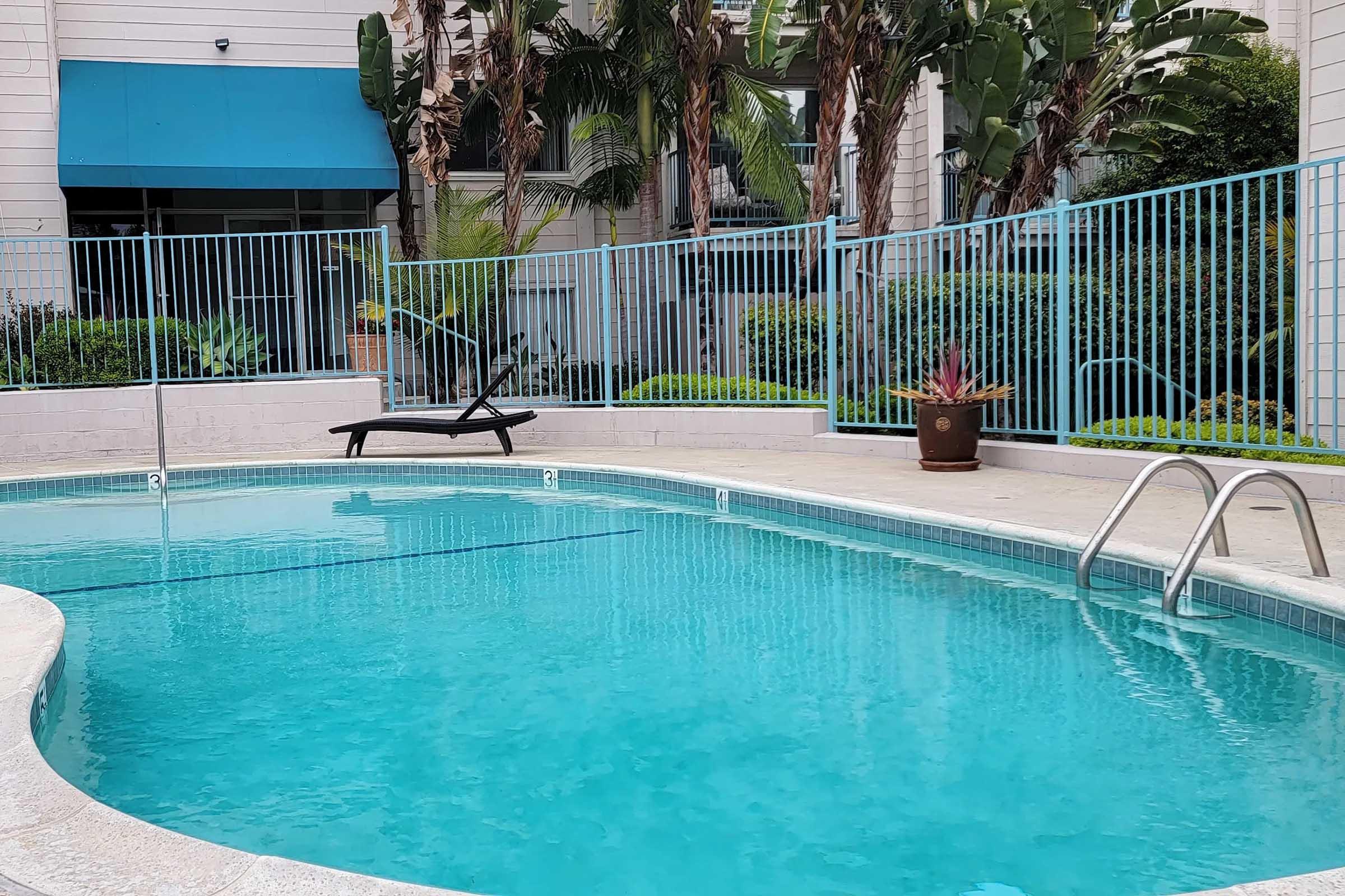 A clear blue swimming pool surrounded by a light-colored patio. There is a black lounge chair positioned beside the pool, while tropical plants and palm trees are visible in the background. A blue building facade with large windows can be seen, and the area is enclosed by a light blue fence.