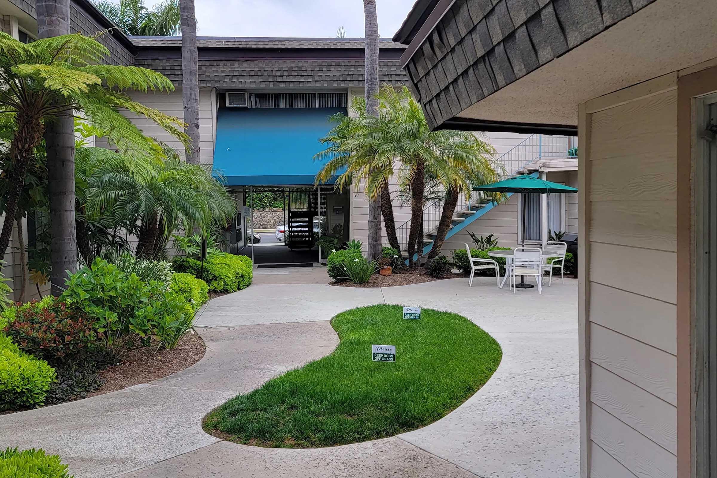 View of a landscaped courtyard featuring lush green plants, palm trees, and a curved pathway. In the background, there is a building with a bright blue awning and outdoor seating areas, including white chairs and tables under umbrellas. The scene is peaceful and inviting.