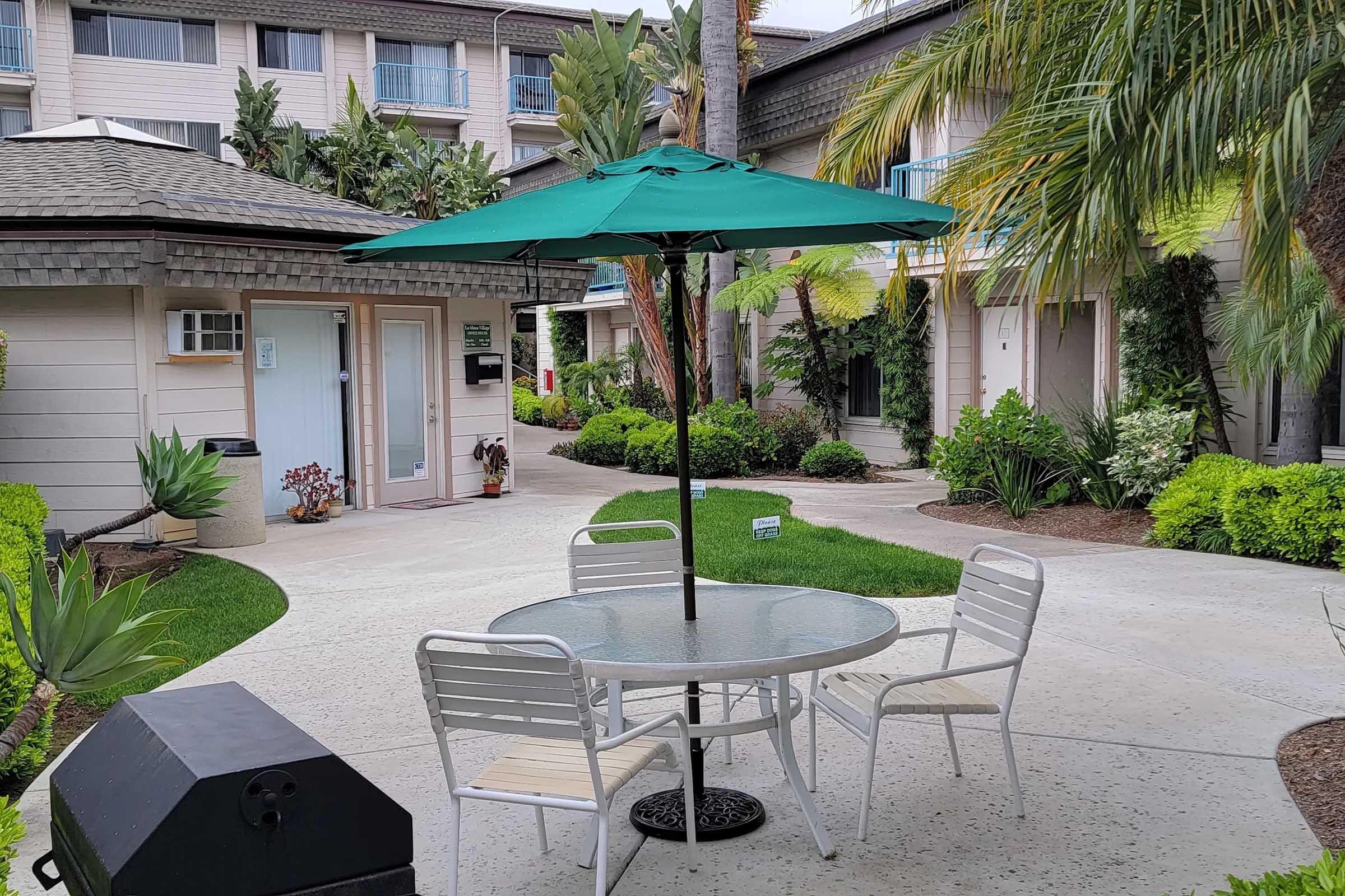 A circular table with four white chairs and a green umbrella is set up in a landscaped courtyard. Lush greenery and palm trees surround the area, with a view of a building in the background. The path is made of light-colored concrete, and a grill is visible nearby.