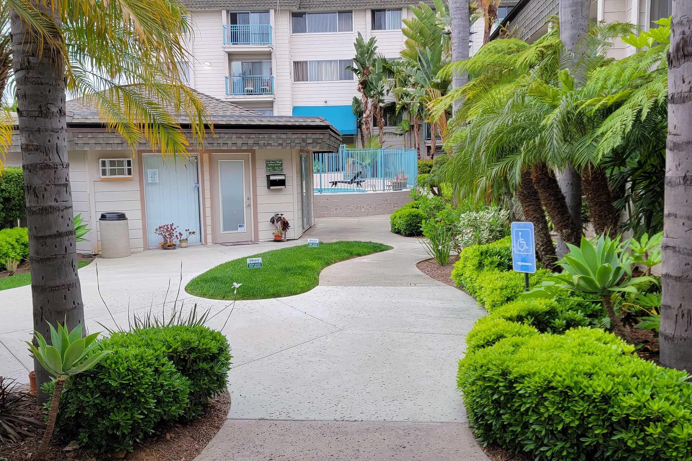 A lush green pathway winds through a tropical garden, featuring palm trees and manicured shrubs. On one side, a swimming pool area is visible, along with a light-colored building. Signage indicating paths is placed throughout, creating a welcoming atmosphere.