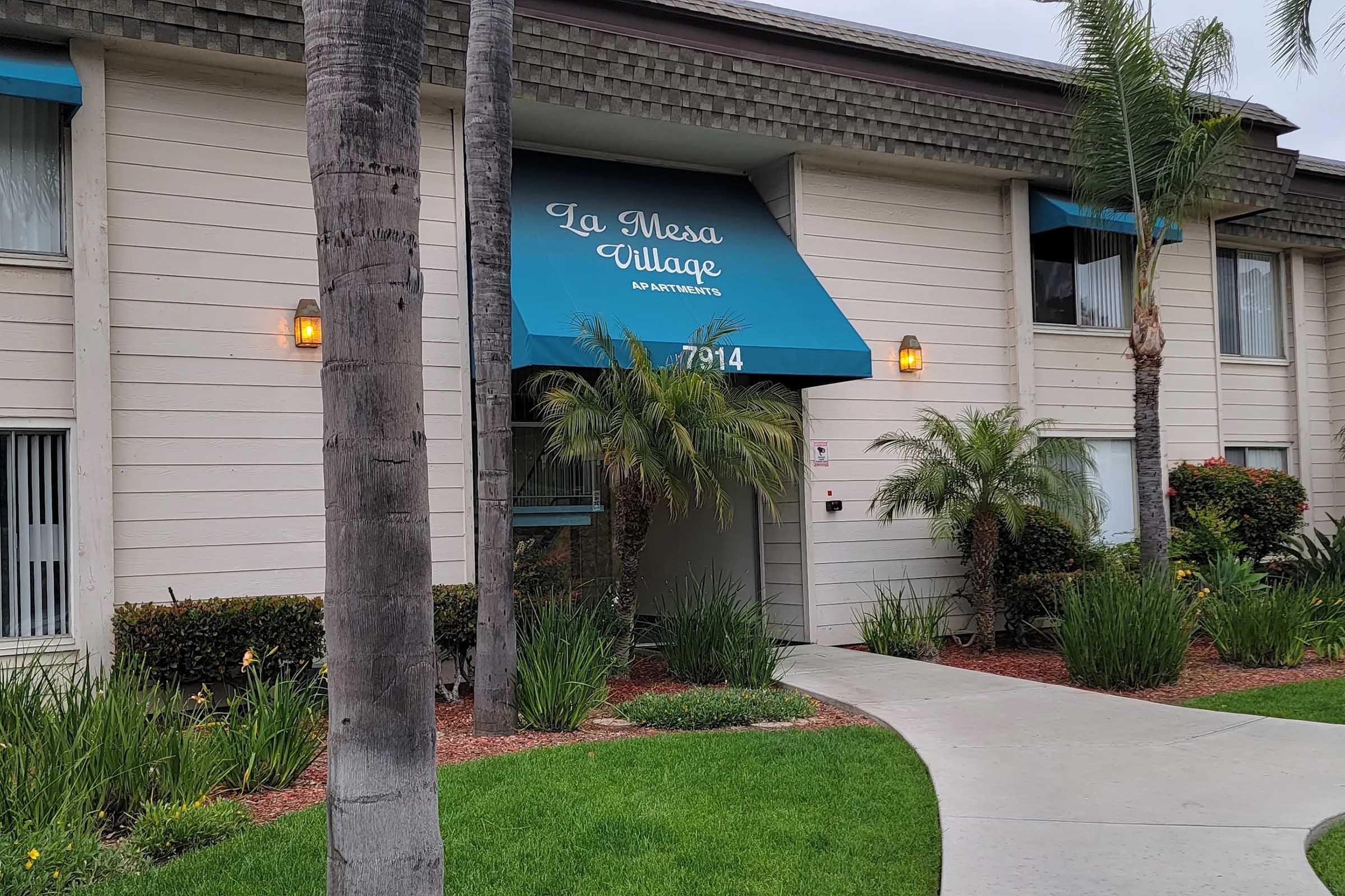 A residential building with a sign reading "La Mesa Village" above the entrance. The building features light-colored siding and blue accents, with palm trees and greenery surrounding the entryway. A curved pathway leads to the entrance, enhancing the inviting atmosphere.