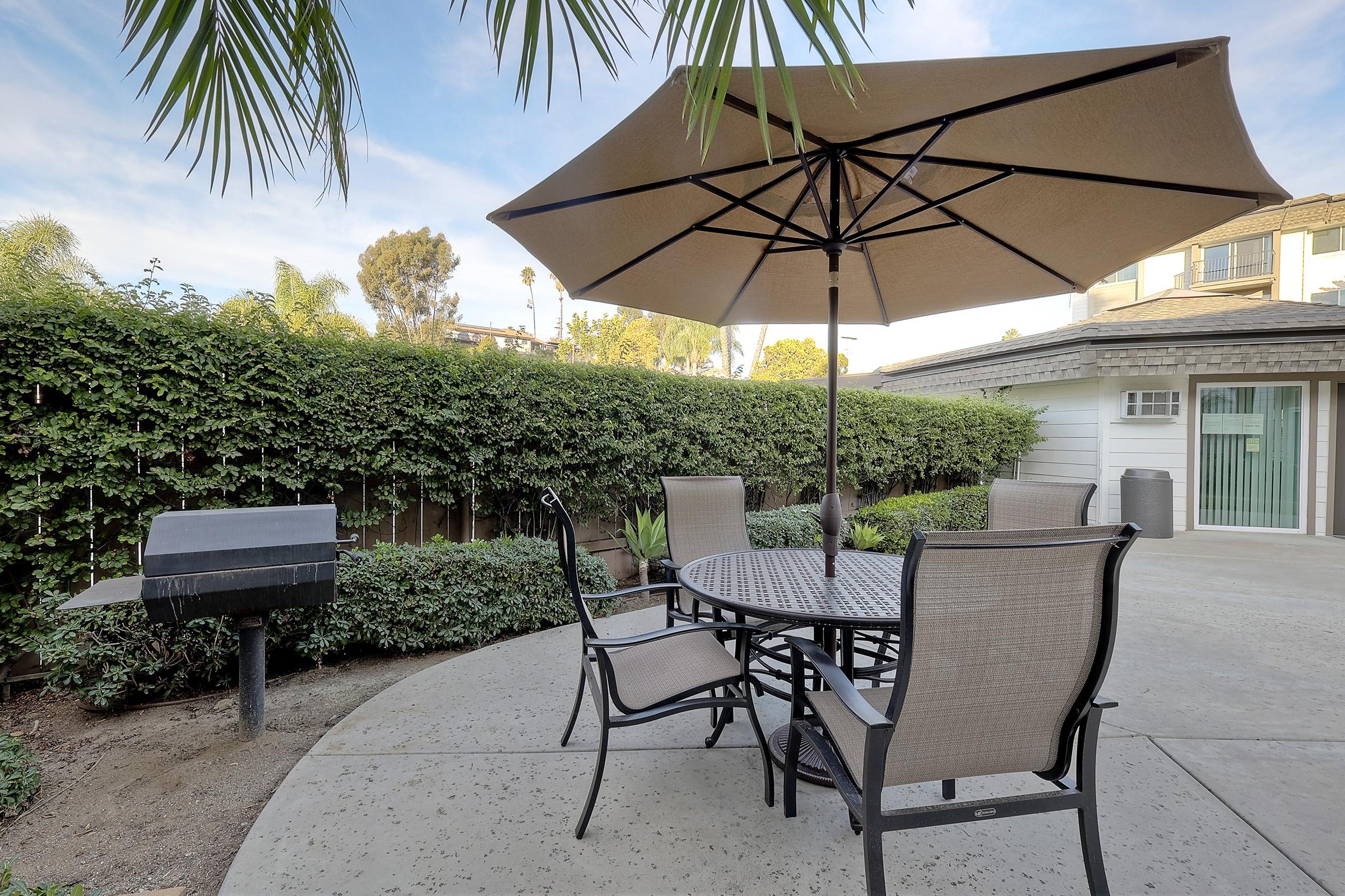 A cozy outdoor patio scene featuring a round table with four chairs underneath a large umbrella. A charcoal grill is positioned nearby, surrounded by lush greenery and well-kept plants. The background includes a residential structure and trees, suggesting a relaxing backyard atmosphere.