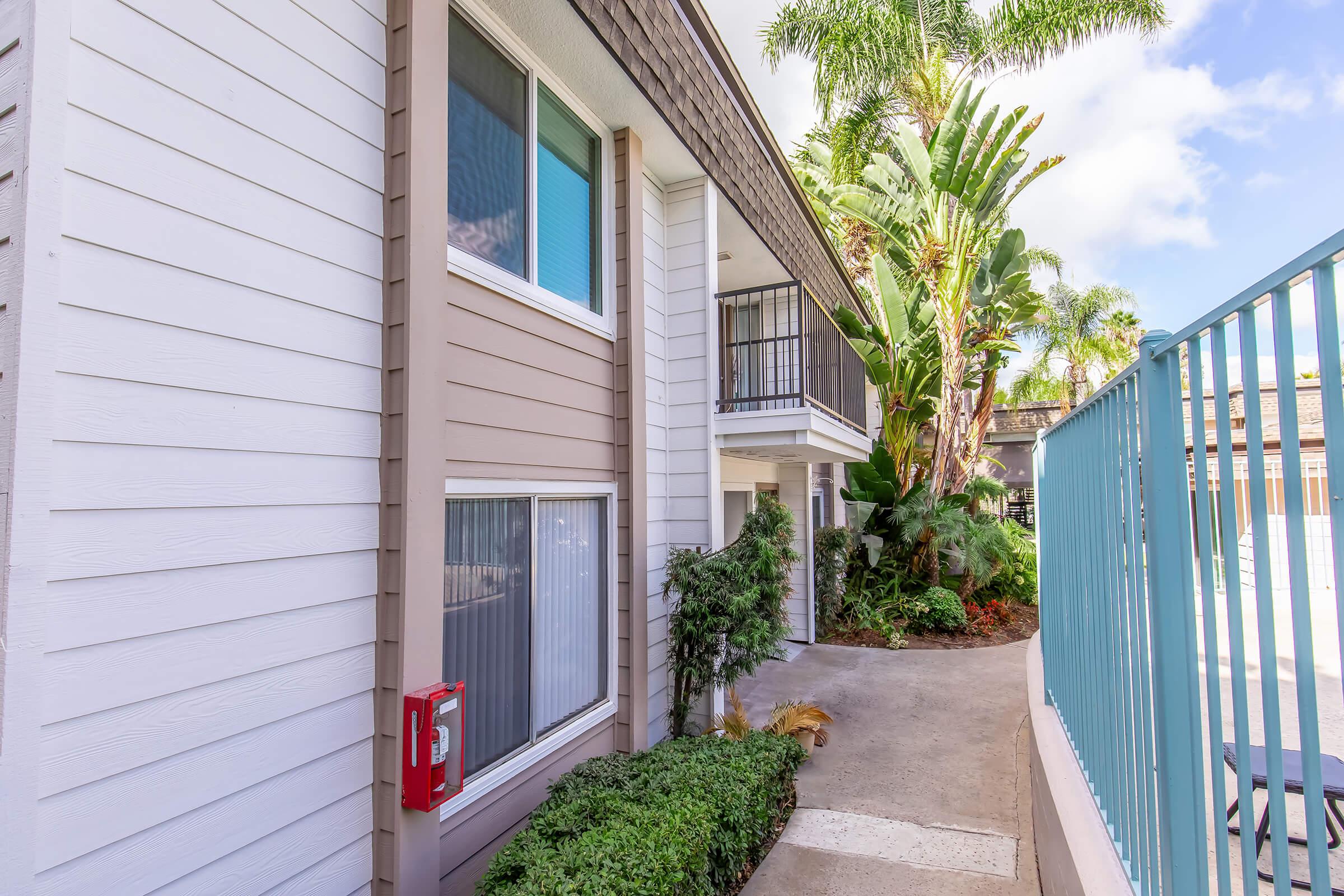 A view of a modern apartment building with light-colored siding. The entrance is flanked by lush greenery and tropical plants. A fire extinguisher is mounted on the wall, and a pathway leads to a balcony on the second floor. Bright blue skies and palm trees can be seen in the background, creating a welcoming atmosphere.