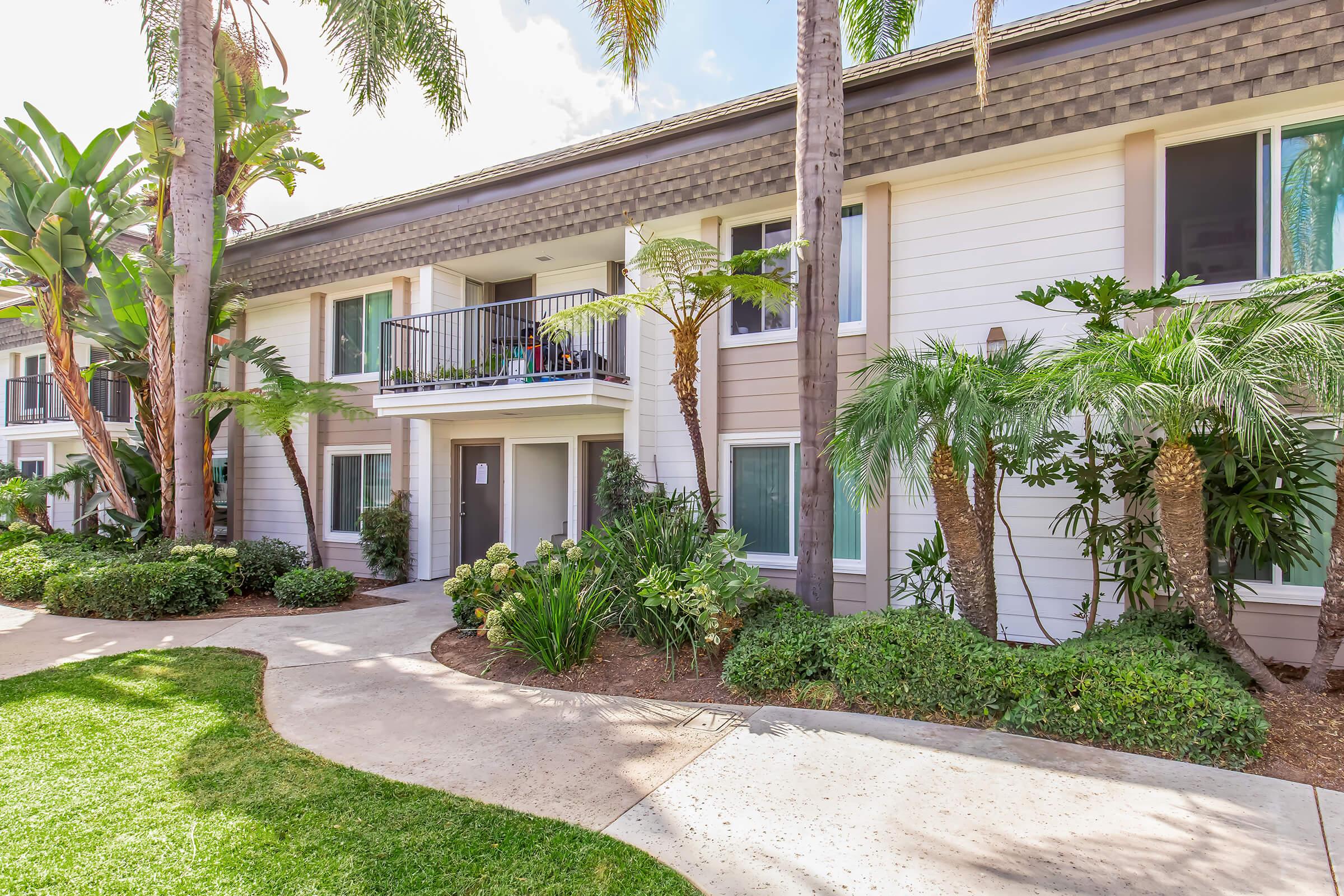A well-maintained outdoor area of a modern apartment building, featuring lush green landscaping with palm trees and shrubs. The building has a light-colored exterior, balconies with plants, and a courtyard pathway leading through the greenery. Bright sunlight illuminates the scene, creating a welcoming atmosphere.