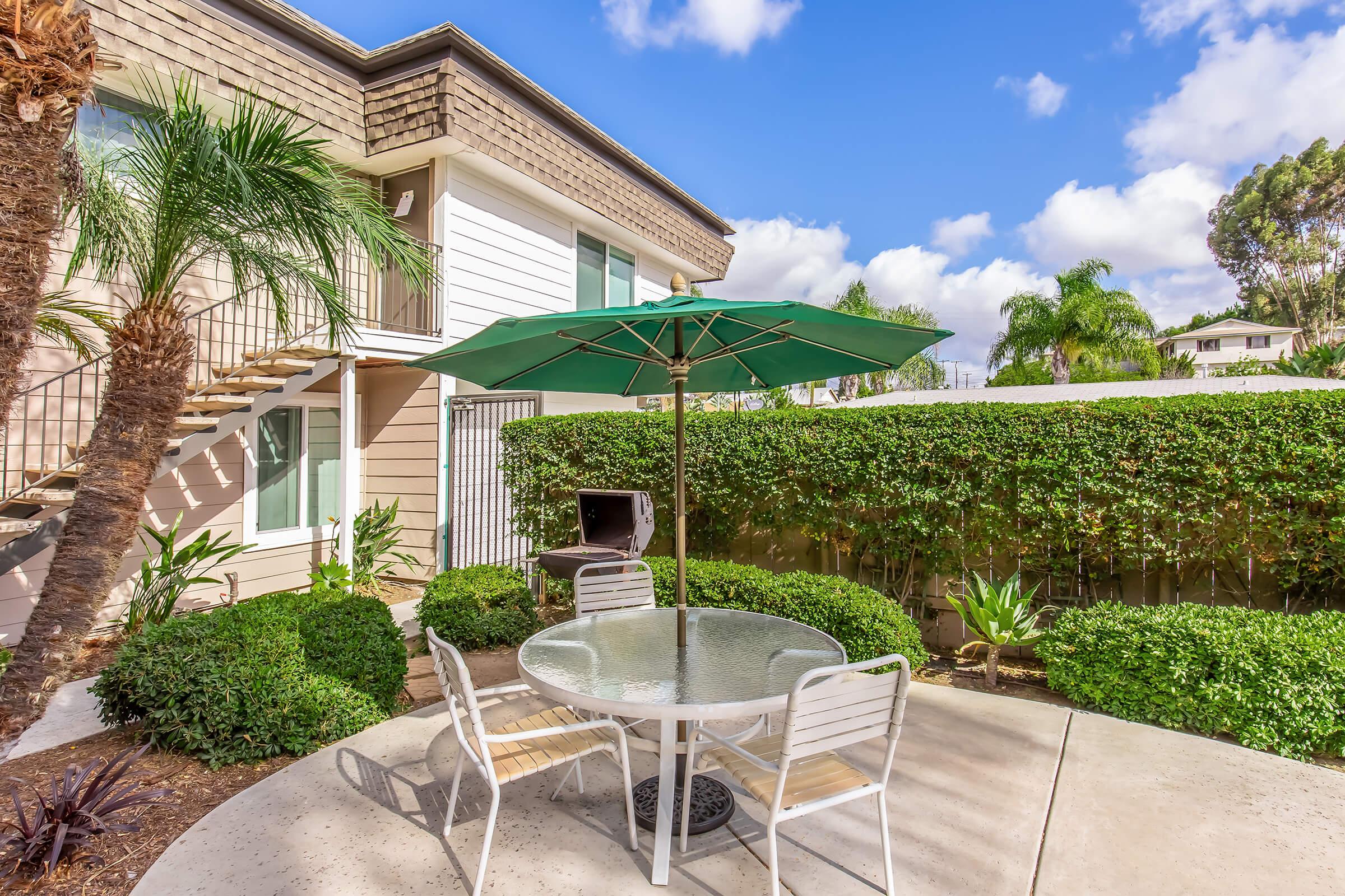 A well-landscaped outdoor area featuring a round glass table with four white chairs, a green umbrella for shade, and a barbecue grill nearby. Lush greenery and palm trees surround the space, with a residential building in the background under a clear blue sky.