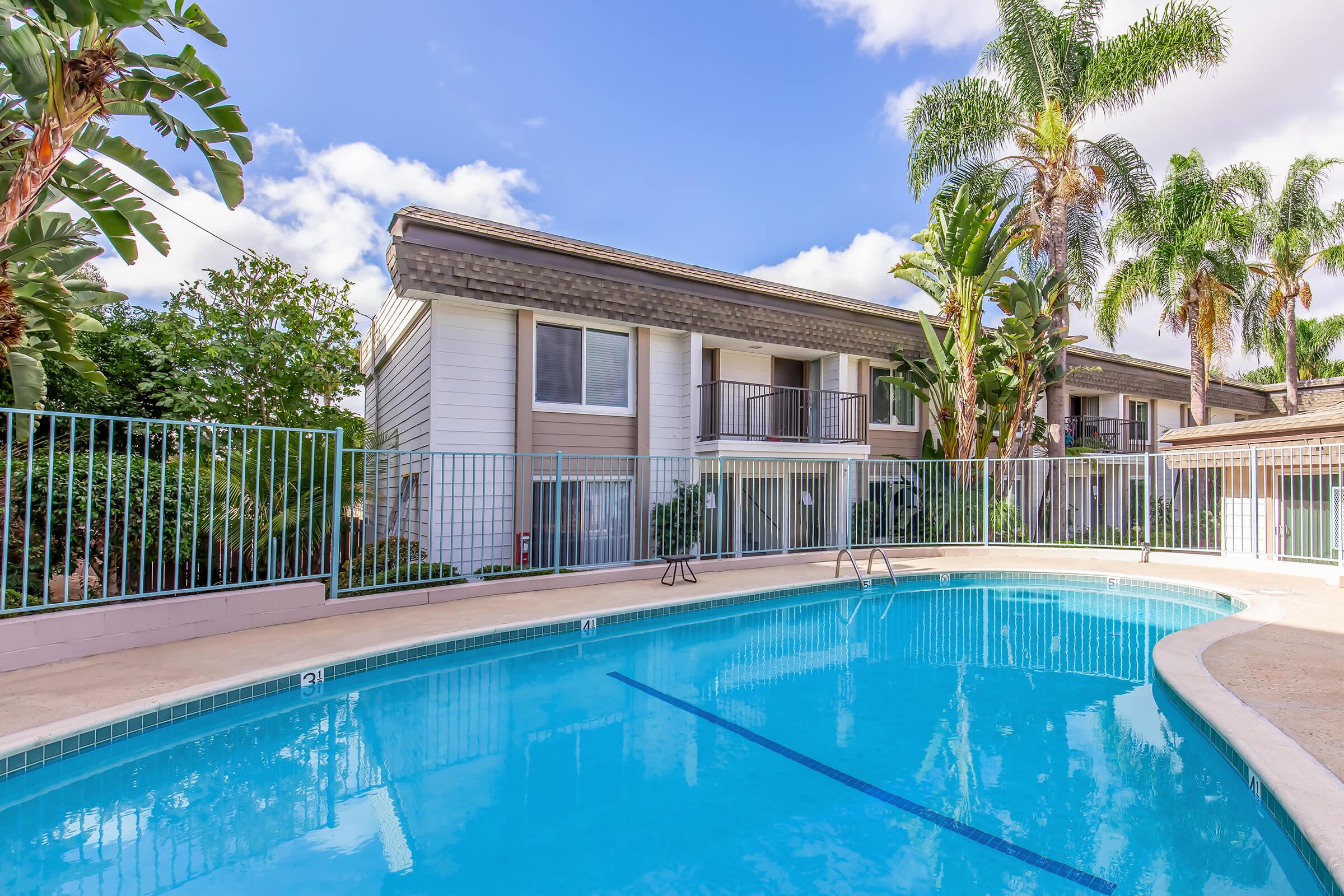 A clear blue swimming pool surrounded by a fenced area, with lush greenery and palm trees in the background. In the distance, a multi-story building features balconies and large windows, under a bright sky with scattered clouds.