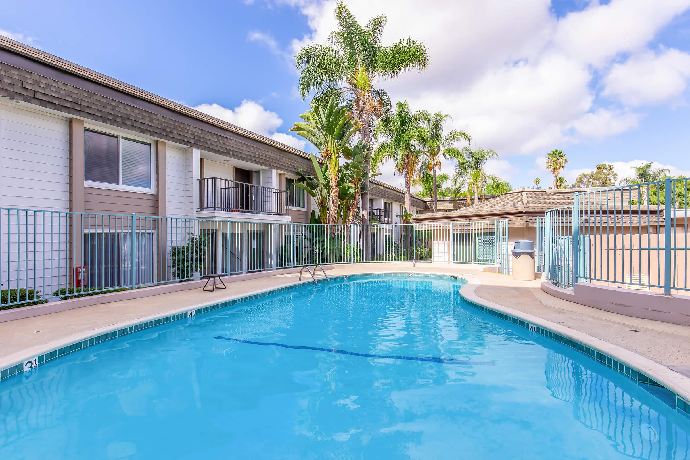 A bright blue swimming pool surrounded by a fenced area, with palm trees and greenery in the background. The pool area is flanked by two multi-story residential buildings, showcasing a serene outdoor space under partly cloudy skies.