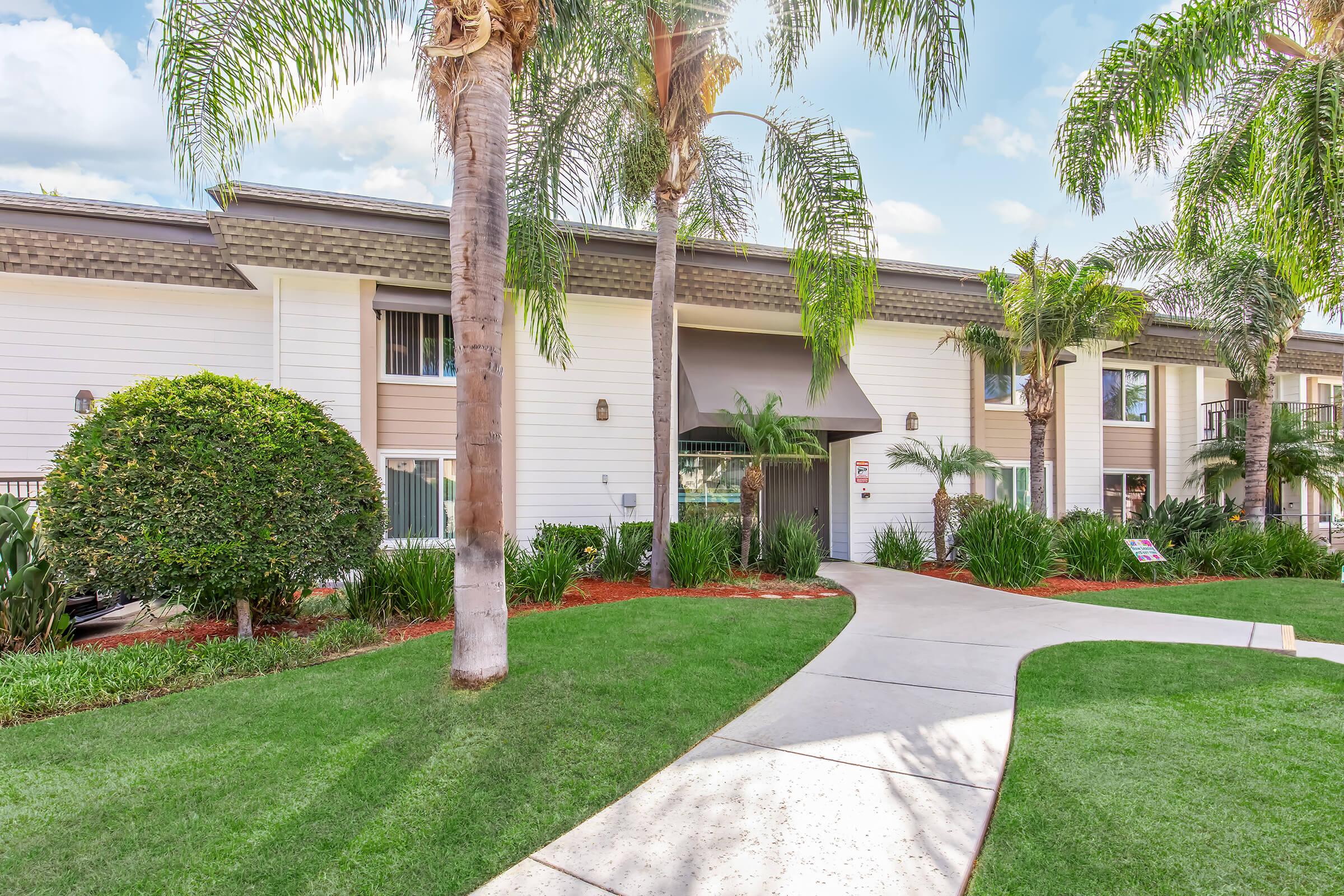 A well-maintained apartment complex surrounded by lush green landscaping, featuring palm trees and shrubs. A walkway leads to the entrance, with bright flowers and manicured lawns visible. The building has a modern design with large windows and a welcoming atmosphere.