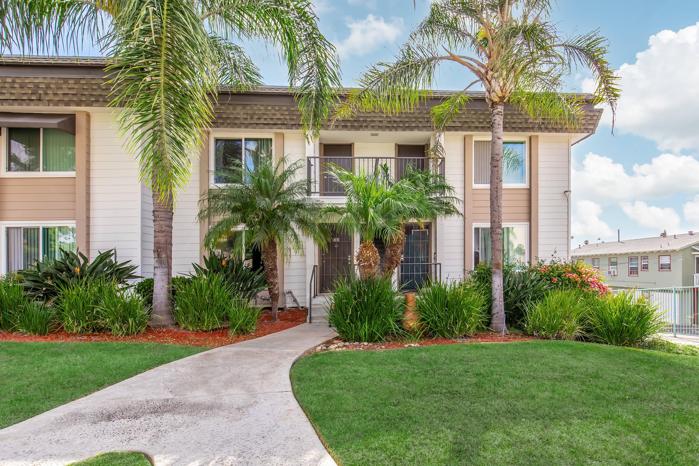 A well-maintained exterior of a multi-unit residential building surrounded by lush greenery, palm trees, and colorful plants. The pathway leads to the entrance, and the sky is partly cloudy, creating a bright and inviting atmosphere.