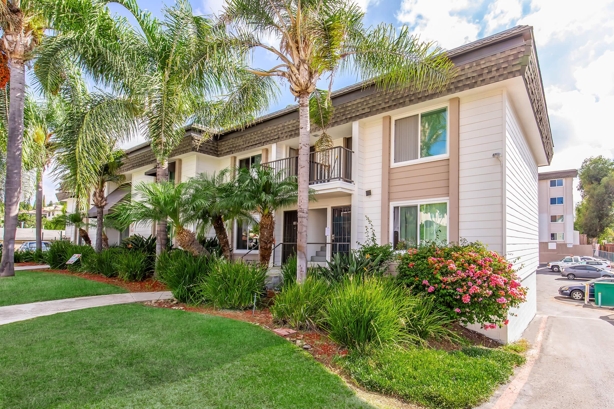 A landscaped view of an apartment complex featuring palm trees, shrubs, and colorful flowers. The building has a mix of white and brown siding with balconies. The surrounding lawn is well-maintained, creating a welcoming outdoor environment.
