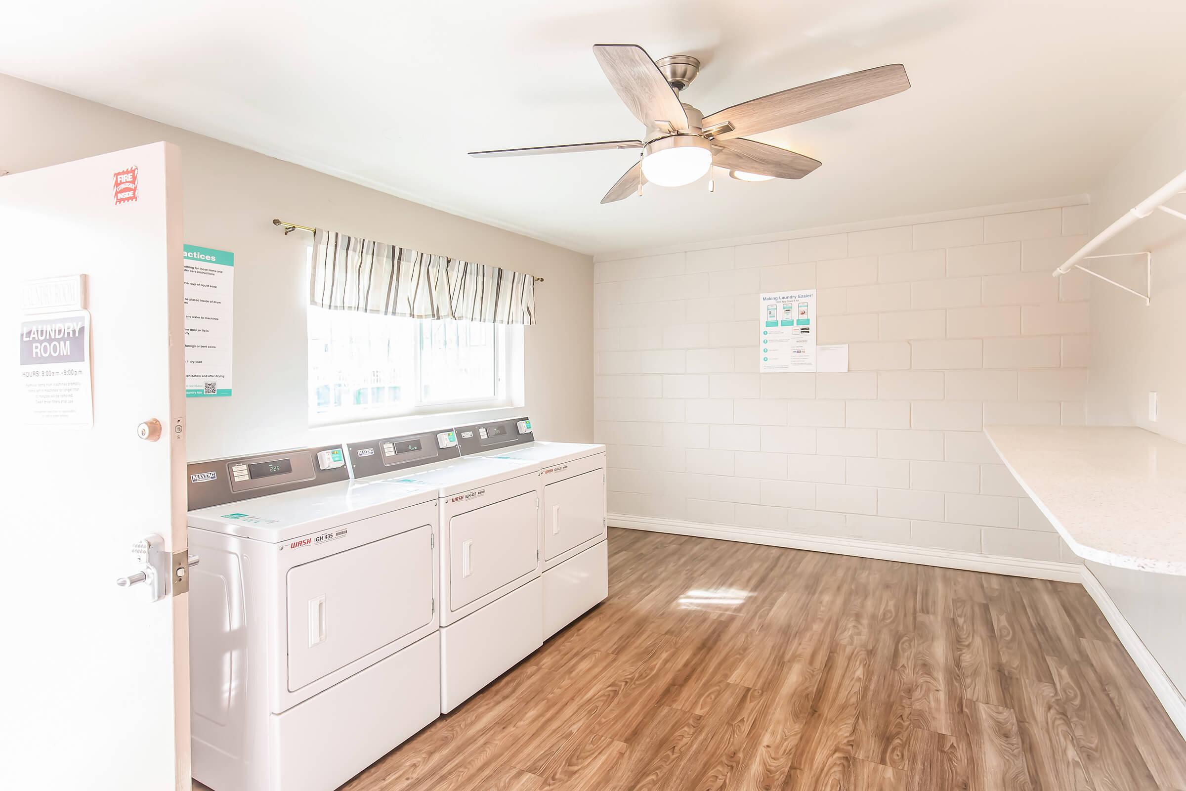 Bright and spacious laundry room featuring two white washing machines and a dryer, a ceiling fan, a large window with light curtains, and open shelving. The room has a clean, modern design with wood-like flooring and informational posters on the walls.