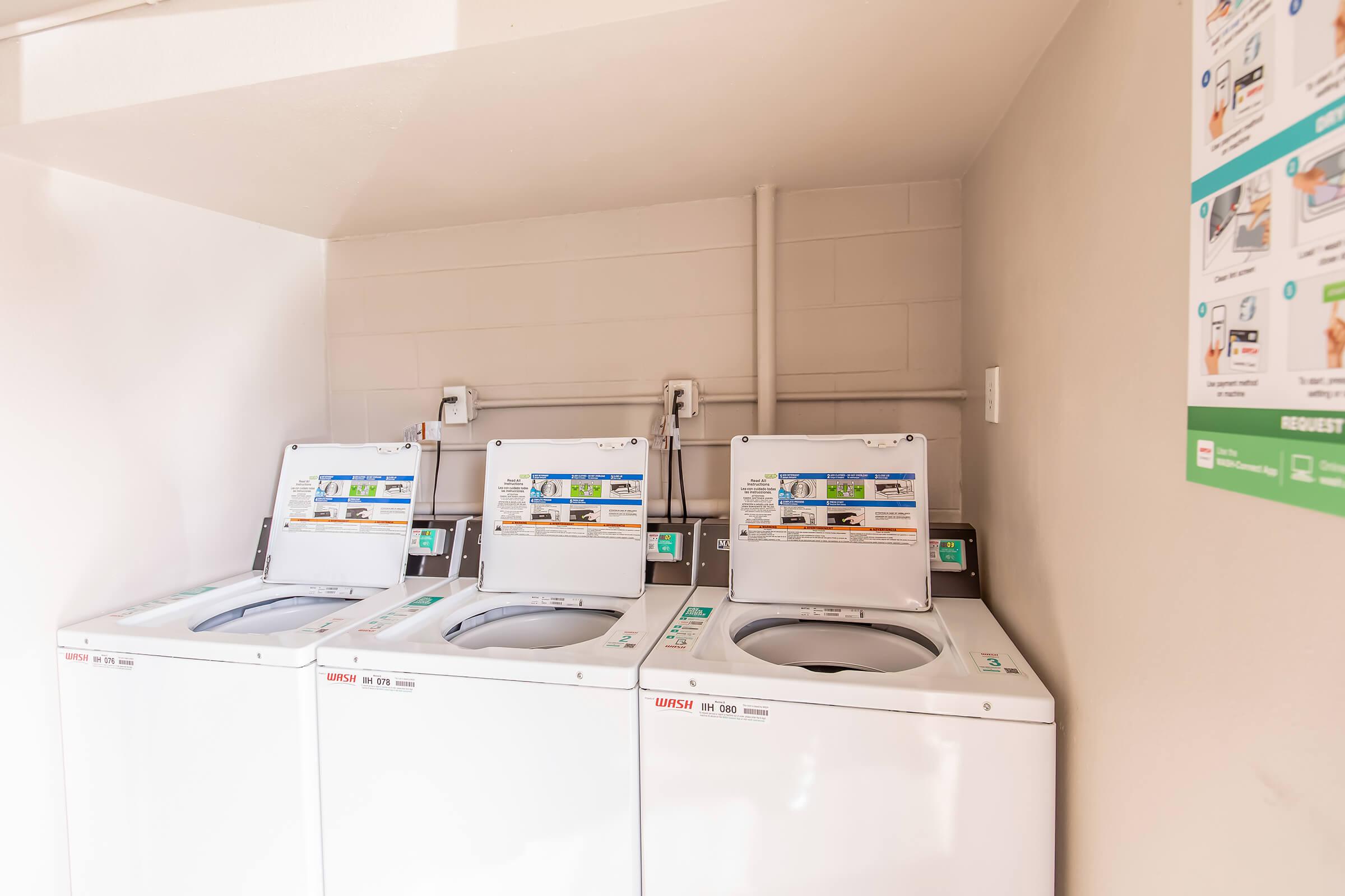 A row of three white washing machines in a clean, well-lit laundry room. The machines are equipped with user-friendly interfaces, and informational posters are visible on the wall nearby, describing laundry procedures and maintenance requests.