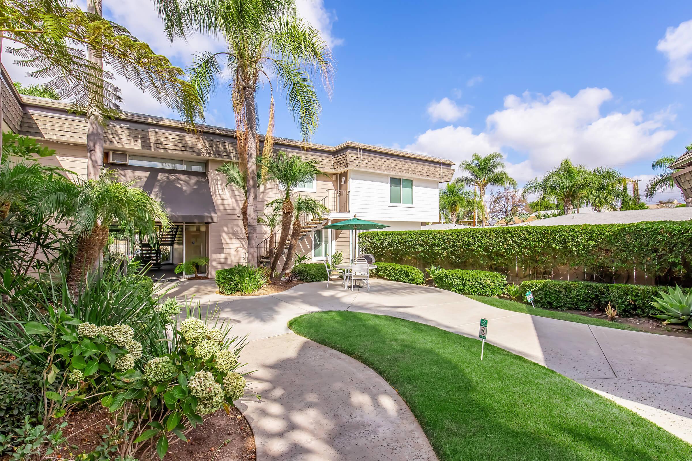 A landscaped courtyard featuring lush greenery, palm trees, and a light-colored building. There is outdoor seating with a green umbrella on a stone pathway leading to a well-maintained lawn under a bright blue sky with fluffy clouds.