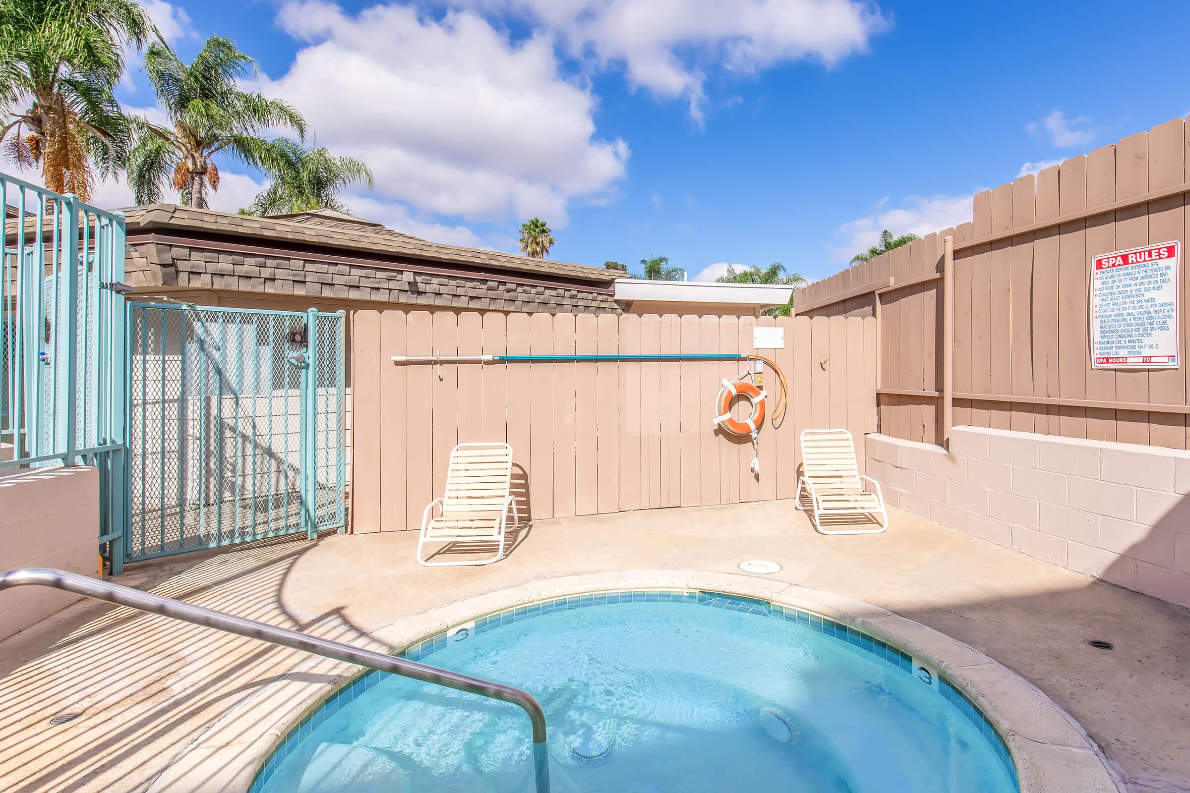 A small, private hot tub area featuring a circular hot tub surrounded by a beige patio. Two lounge chairs are placed beside the hot tub. A wooden fence with a safety sign and a life ring hangs on the wall. Palm trees and a partly cloudy sky are visible in the background.