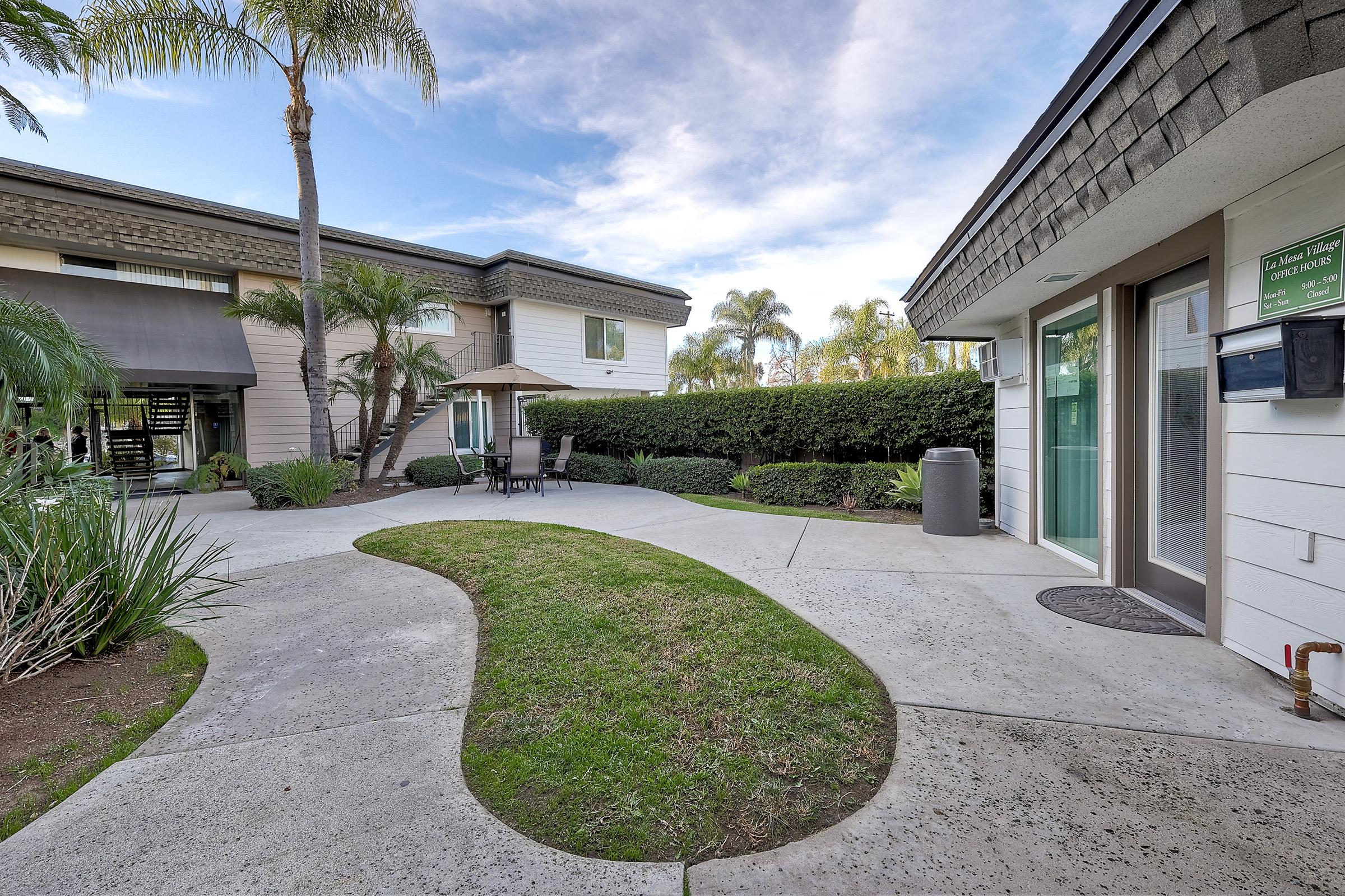 A landscaped outdoor area featuring a curved pathway, green grass, palm trees, and a seating area with chairs under an umbrella. The scene includes two buildings with modern architecture, plants, and a mailbox near a door. The sky is partly cloudy, creating a pleasant atmosphere.
